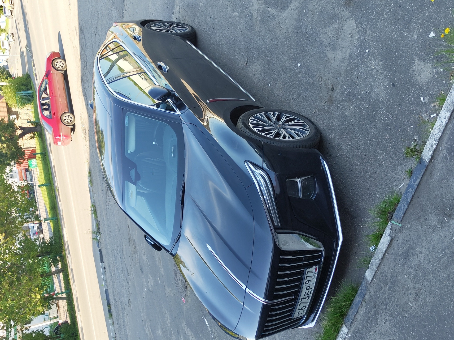 A black sedan is parked on an asphalt road in Zhukovsky, Russia. The car is facing right, and its front grille, headlights, and license plate are visible. The license plate reads "C673EP 977 RUS". A red sedan is driving on the road in the background, also facing right. Trees and buildings are visible in the background. The sun is shining, casting shadows on the ground.
