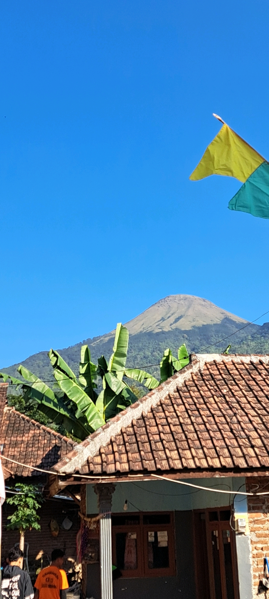 In the city of Sukoreno, Indonesia, a clear blue sky forms the backdrop for a scene featuring a prominent, conical mountain topped with vegetation. In the foreground, a tiled roof of a dwelling extends across the frame. Large, green banana leaves fan out from behind the roof. Two flags, one yellow and one turquoise, are visible in the upper right portion of the image, fluttering against the sky. Below the roof, the exterior of a house is visible, with brickwork and windows. Two young men are partially visible in the lower left of the frame, one wearing a black shirt with white lettering and the other an orange shirt.