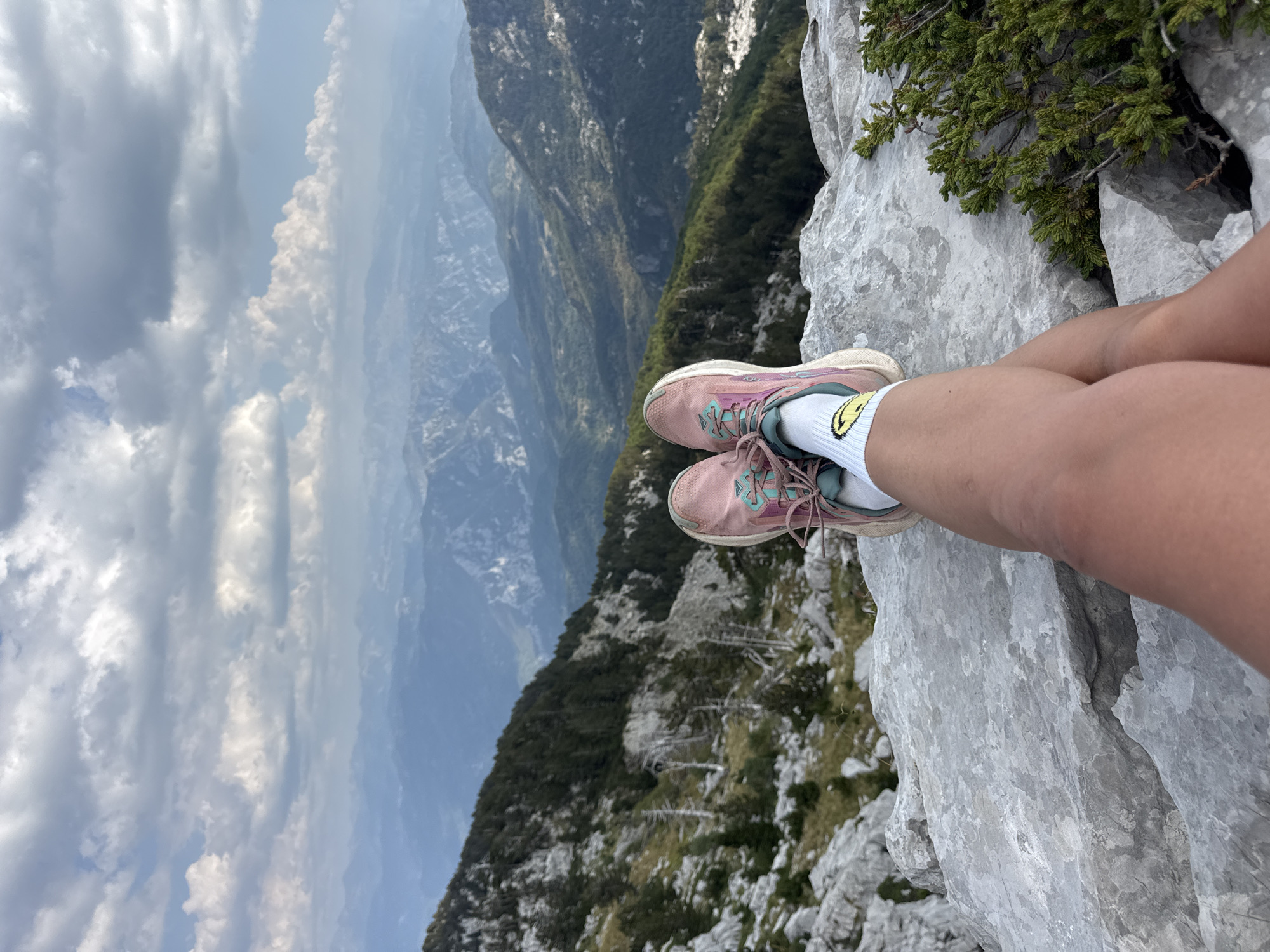 A person's legs, wearing pink athletic shoes and white socks with a yellow smiley face graphic, are crossed while sitting on a rocky outcrop. The background is a vast mountainous landscape with layers of hazy blue mountains receding into the distance under a cloudy sky. The immediate foreground consists of large, light-grey rocks with sparse green vegetation. The terrain behind the person slopes downwards, covered in dark green coniferous trees and rocky, sparsely vegetated areas. The scene appears to be located in a high-altitude, natural environment.