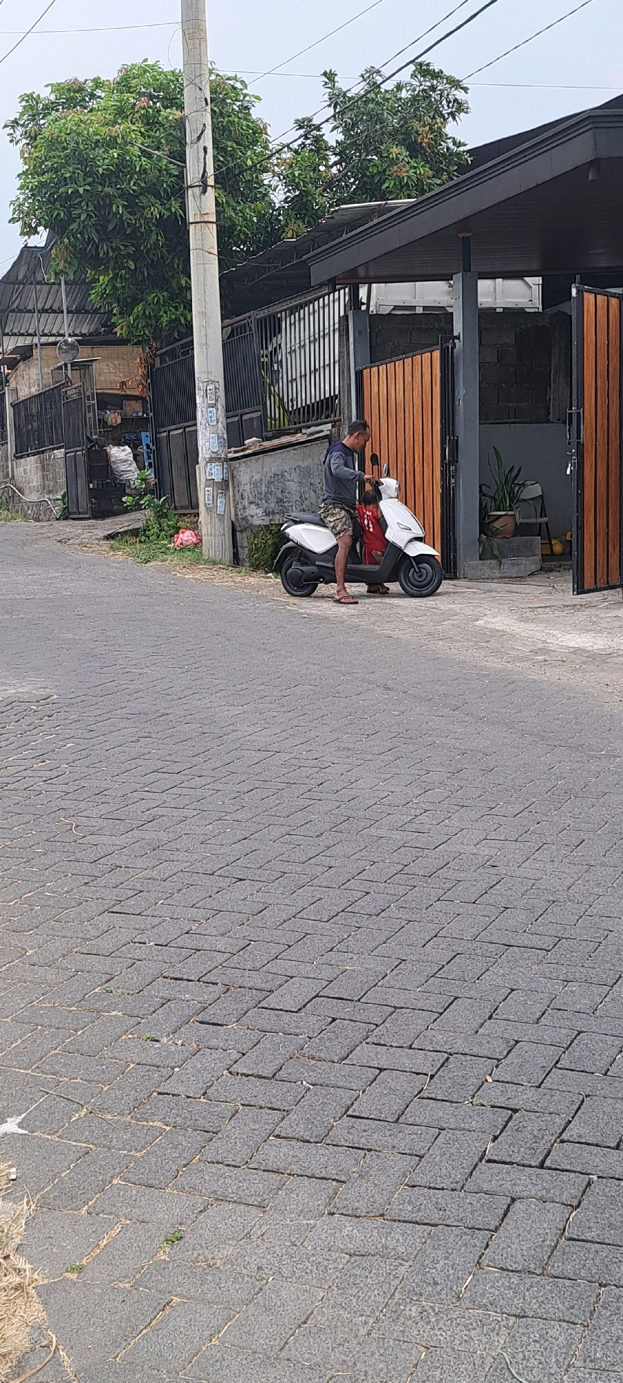 A man is sitting on a white scooter in the street of Ketanireng, Indonesia. He is wearing a gray long-sleeved shirt and patterned shorts. The scooter has a red bag on the seat. The street is paved with interlocking gray stones. To the left of the scooter, there is a concrete utility pole with wires. Behind the scooter, there is a building with a dark roof and wooden doors. There are trees and fences in the background.