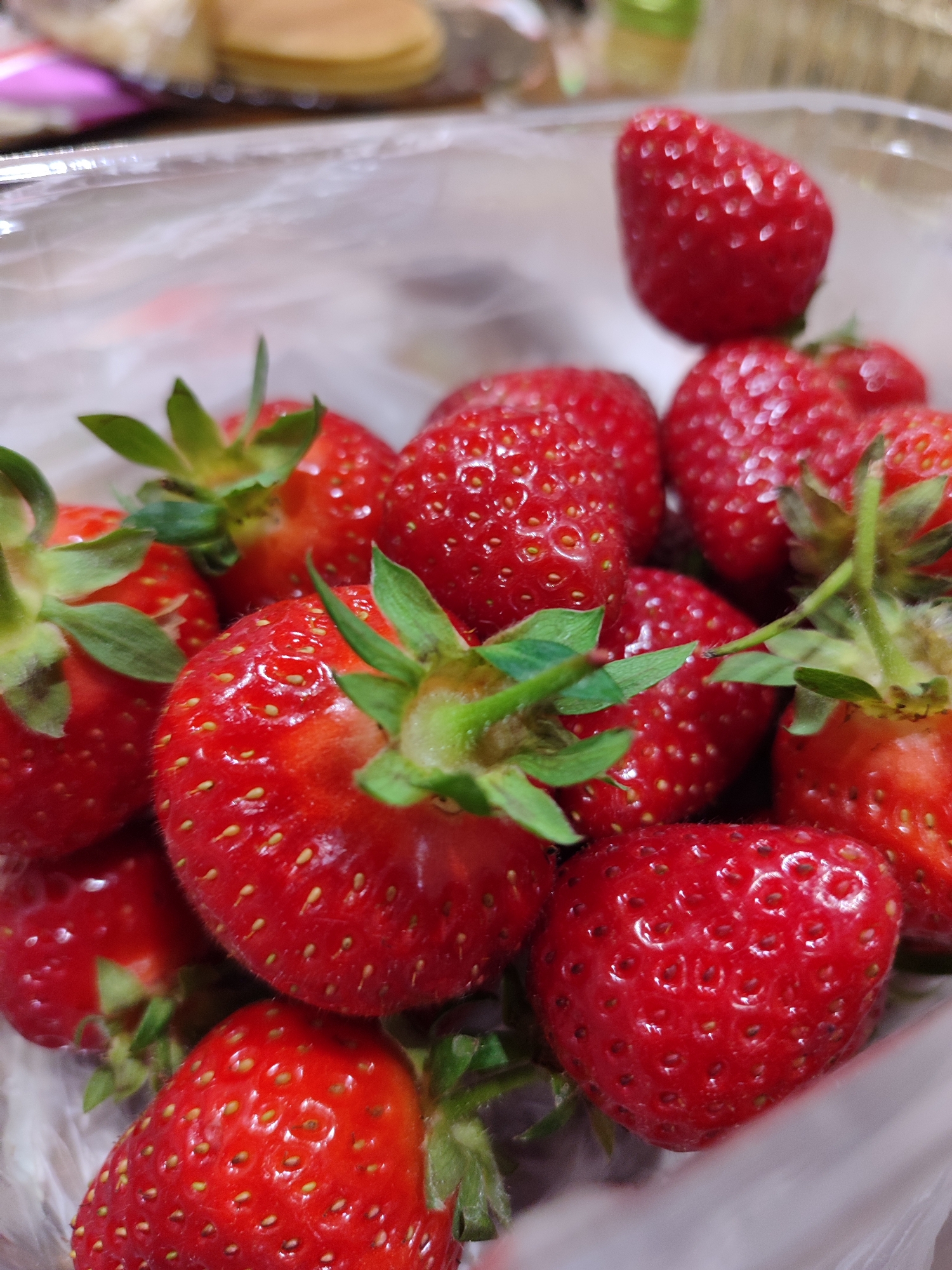 The image shows a close-up of a container of fresh strawberries. The strawberries are red and glossy, with green leafy stems. The container appears to be plastic. The background is blurry, but what looks like pancakes or other food is visible. The location is not identifiable from the image. No people, actions, or interactions are depicted.