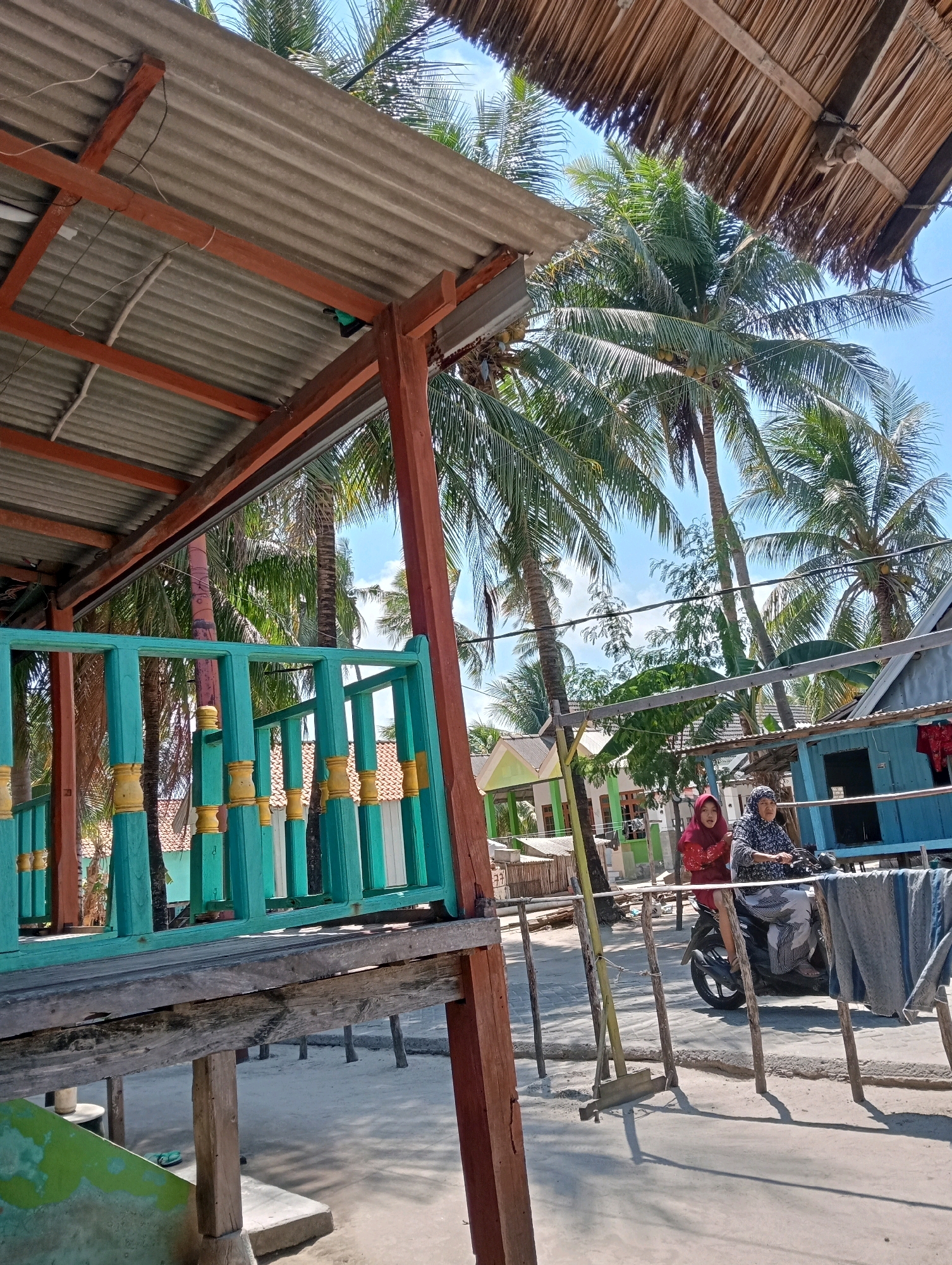 In Pagerungan Kecil, Indonesia, two individuals are captured on a motorcycle on a sandy road, beneath the shade of palm trees and rustic structures. A young person wearing a red hijab is seated in front, while an older person in a patterned hijab drives. Their motorcycle is parked near a wooden structure with a teal railing. Behind them, a vibrant blue building stands, with laundry hanging on a clothesline. Further in the background, more houses and lush palm trees under a clear blue sky are visible. The scene suggests a warm, tropical environment, possibly a rural village setting.