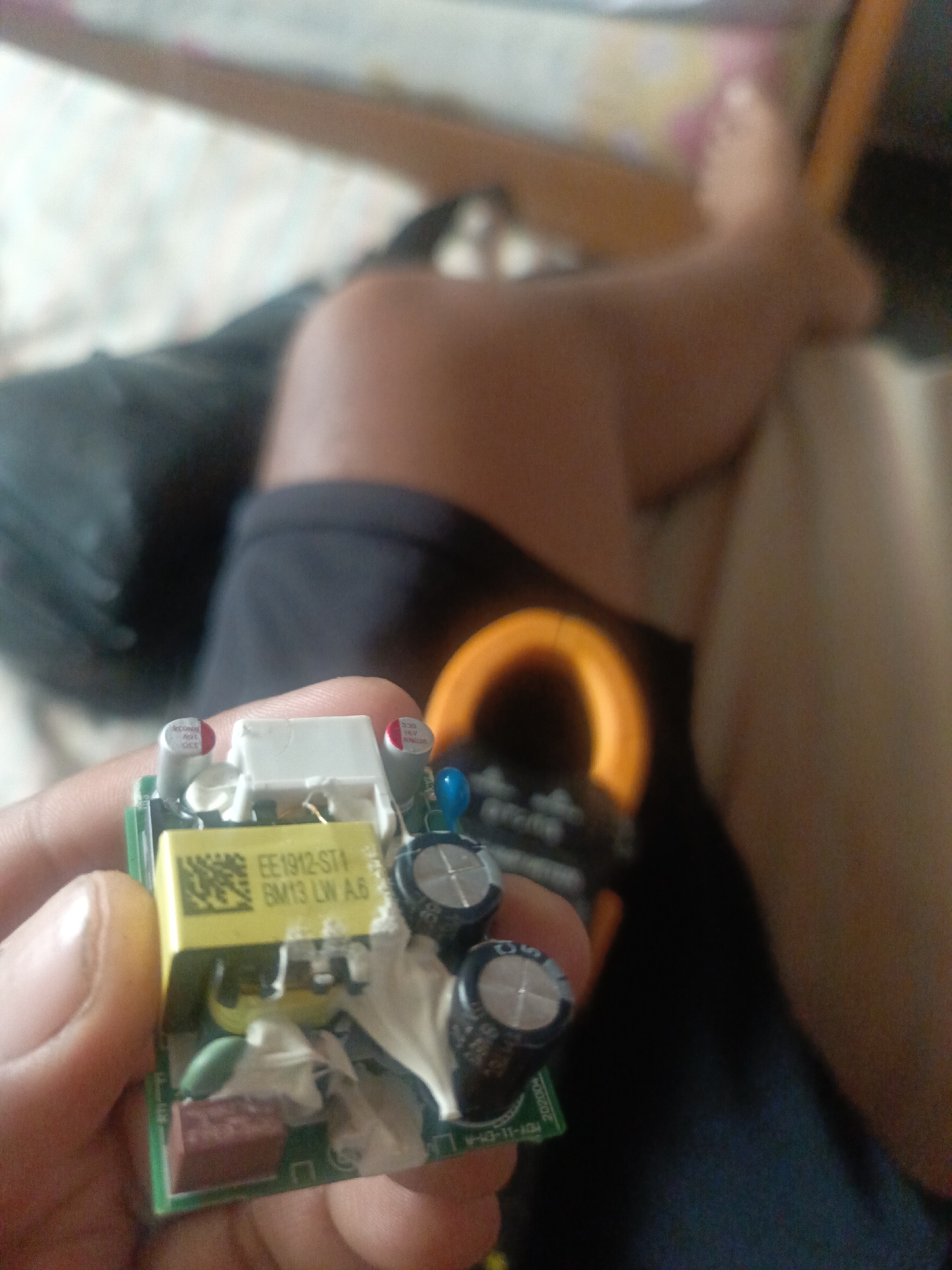 Here's a detailed description of the image:
**Scene:** An indoor setting with a person holding a small circuit board in their hand. The background is blurry, but it appears the person is sitting, possibly on a bed or sofa.
**People:** One person is visible, holding the circuit board. Their skin tone appears to be medium to dark.
**Subjects:** The primary subject is the circuit board. It's green and contains various electronic components like capacitors, a transformer (yellow), and other smaller parts. The label on the transformer reads "EE1912-ST1 BM13 LW A.6". The board also has the marking "M-43-11-AGH 202004" printed on it.
**Objects:**
* Circuit board
* Capacitors
* Transformer
* A clamp meter is partially visible in the background behind the circuit board
**Actions:** The person is holding and presenting the circuit board for viewing.
**Environment:** The environment is indistinct due to the blur.
**Location:** The location is not discernible based on the provided image. I am unable to determine if the image was taken in Juan Griego, Venezuela, or anywhere else.