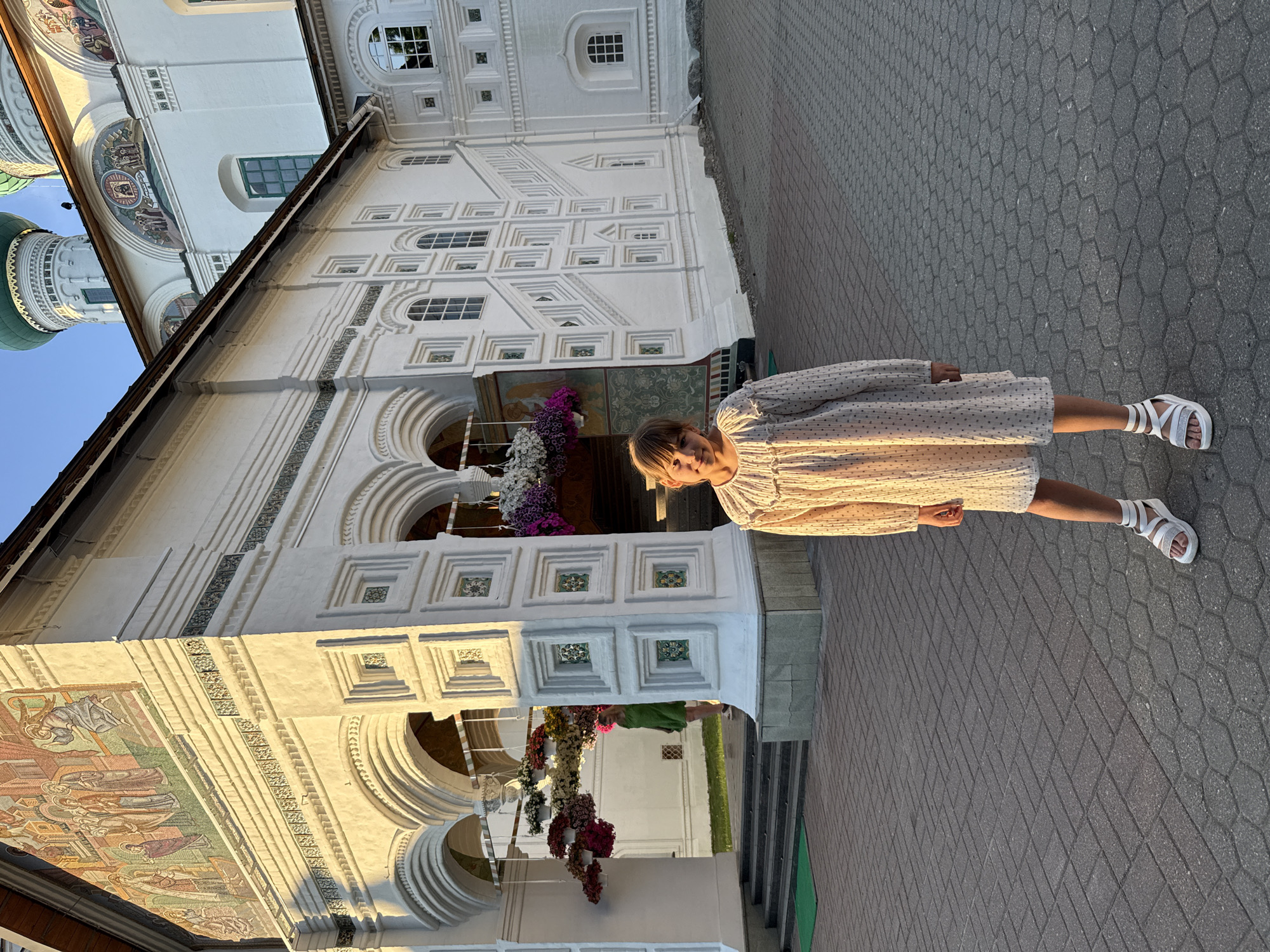 The image displays a young girl, approximately 7-10 years old, standing in the foreground, facing the camera with a slight smile. She has light brown hair with bangs and is dressed in a long-sleeved, cream-colored dress featuring small dark polka dots and ruffled details around the neckline and cuffs. She wears white platform sandals over white socks.
Behind the girl is a large, multi-story white building, characterized by intricate architectural details including arches, pilasters, and decorative square patterns. The building exhibits multiple colorful mosaic murals; a prominent, multi-figure mosaic is on the upper left section of the facade, and a smaller mosaic is visible on a higher structure. Two green onion domes rise above the white walls against a clear blue sky on the upper right. Several hanging flower baskets containing white, pink, and purple blooms are suspended near arched entrances on the left side of the building. Another person, visible from the back and side in a green dress, is ascending a set of steps into one of these arched doorways on the left.
The ground in the foreground is paved with grey hexagonal interlocking pavers. The scene is illuminated by warm, low-angle sunlight, indicating late afternoon or early evening. This building is located in Kuznechikha, Russia.