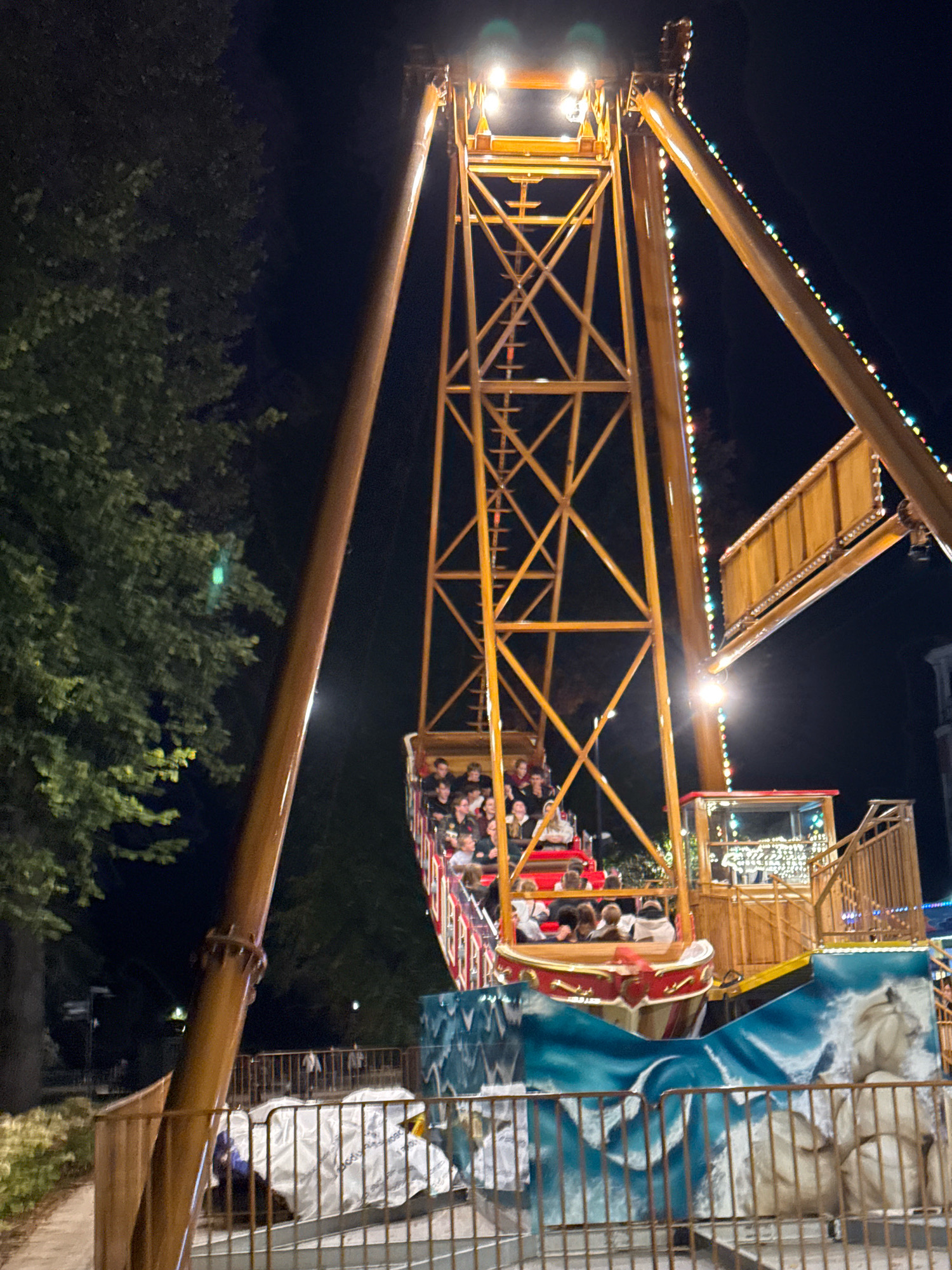 The image depicts a pirate ship amusement ride at night in an outdoor setting, likely a park or fairground, in the city of Tver, Russia. The ride is illuminated by strings of colorful lights and spotlights. A boat-like structure, filled with people, is suspended from a tall, yellow, metal frame. The boat is decorated with red and gold trim and features a painted wave design at its base. Some of the passengers are visible, appearing to be enjoying the ride. The background is dark, with hints of trees and possibly buildings visible. A metal fence encloses the ride area, with some of the ride's machinery covered by white tarps behind it. The overall atmosphere is festive and nocturnal, characteristic of an amusement park at night.
