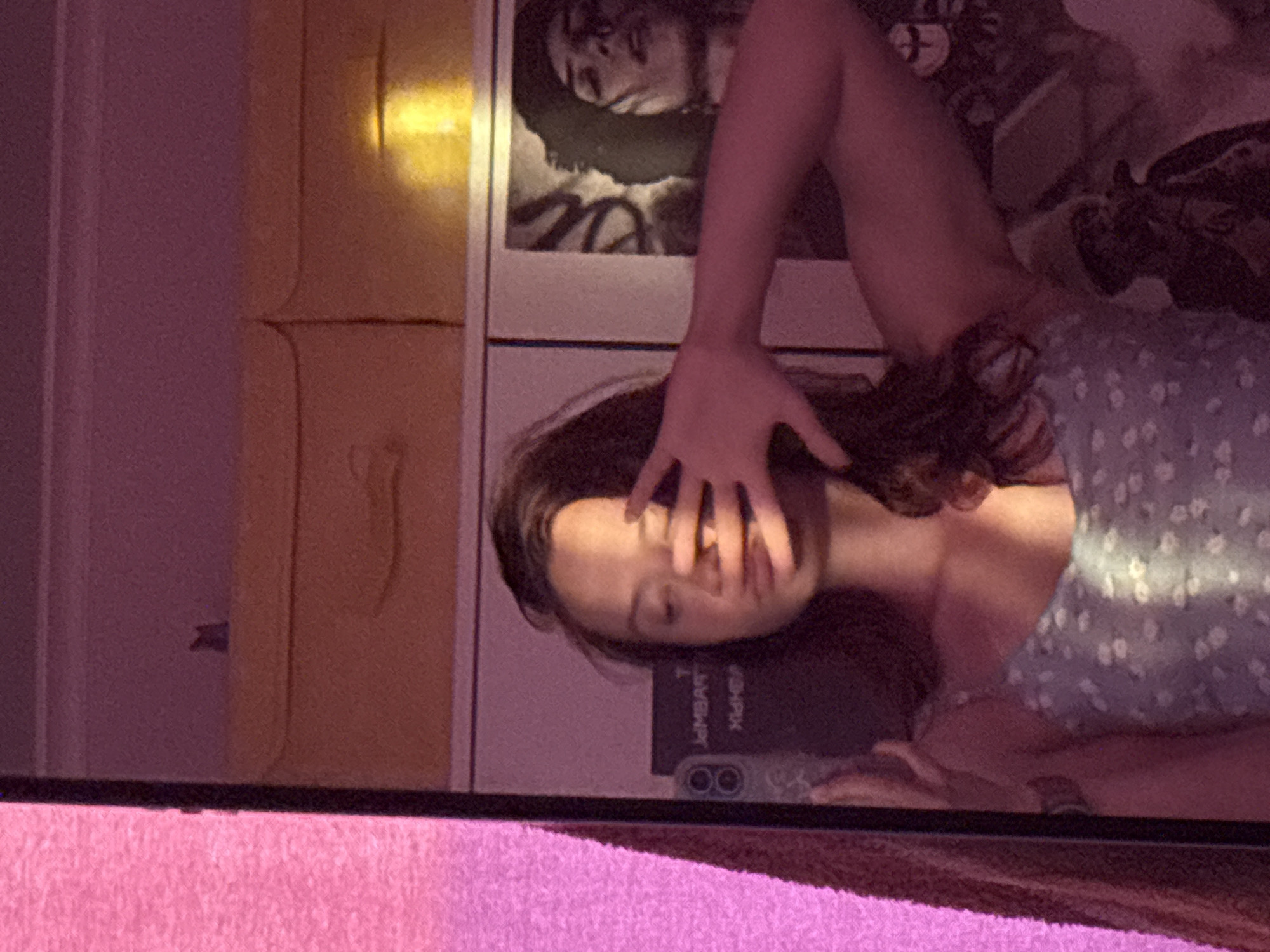 The image shows a reflection in a mirror. A young woman is partially visible, her face obscured by her hand. She is wearing a floral print top. Behind her is a wall with a poster and a yellow storage unit. A cell phone with a case labeled "SHARK T JAMBART" is visible at the bottom. The bottom of the mirror rests on a pink surface. There is no information about the city of Прикамский, Russia.