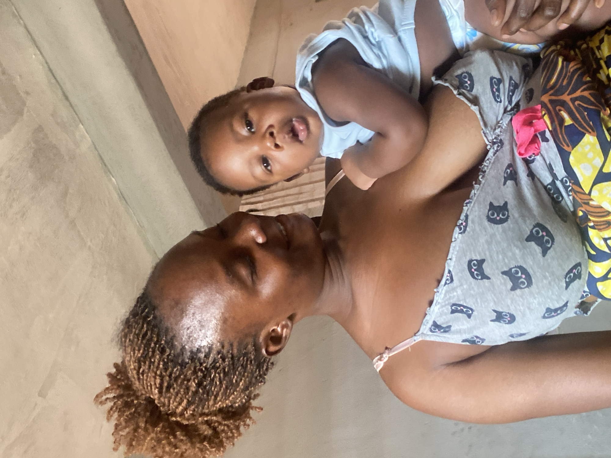 A medium shot shows a dark-skinned woman holding a baby. The woman has light brown braided hair and is wearing a gray tank top with black cat faces printed on it. The baby, swaddled in a white top and diaper, looks toward the camera with its mouth slightly open. The background is a neutral, solid wall, and the lighting appears to be from an overhead source. It is impossible to confirm the location.