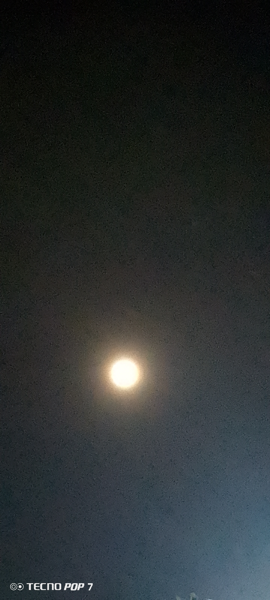 The image shows a bright light source, likely the moon, positioned centrally in a dark sky. The sky appears textured and slightly grainy. At the bottom left corner, the watermark "TECNO POP 7" is visible. The presence of the moon suggests a nighttime environment. The absence of identifiable landmarks and people prevents pinpointing a precise location in Altagracia, Venezuela.