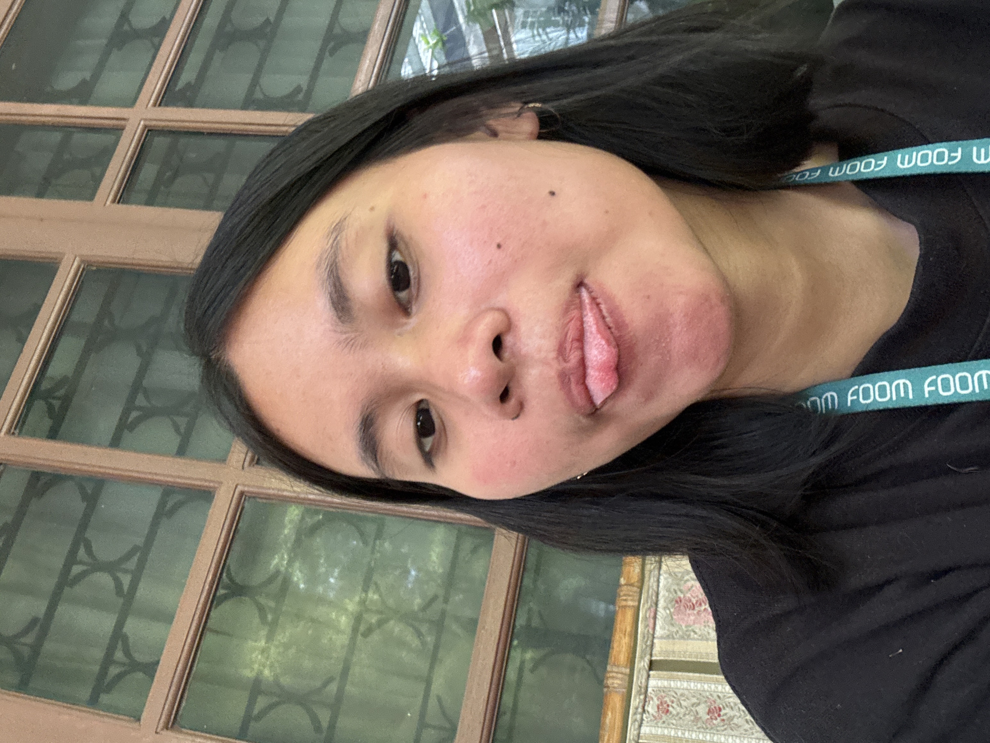 In an indoor setting, a young woman with dark hair and fair skin is captured in a close-up selfie. She is wearing a black t-shirt, and a teal lanyard with white text is visible around her neck. Her face is turned slightly to the right, and she is sticking her tongue out with a playful expression. The background features a window with a wooden frame and decorative ironwork, and behind the window, a hint of greenery suggests an outdoor environment. The room appears to be furnished with a patterned upholstered bench. The overall lighting is natural, casting a soft glow on the woman's face. No specific landmarks of South Jakarta are visible in this image.