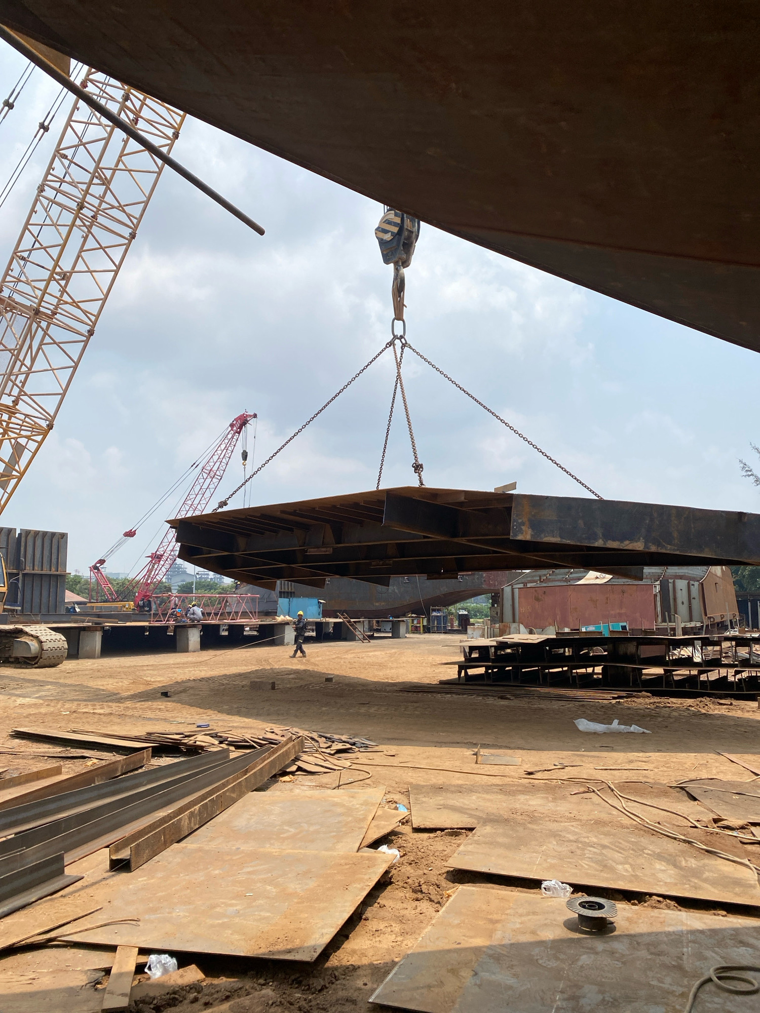 This is an outdoor industrial scene in Batam, Indonesia, likely a shipyard or heavy fabrication facility. A large, flat metal structure, possibly a ship hull section or bridge component, is being lifted by a crane via heavy chains. In the background, a red crane is visible, along with other construction materials and heavy machinery, including a large yellow excavator. Several individuals are present on the ground, some appearing to be workers overseeing the operation. The ground is covered with dirt, metal scraps, and pre-fabricated steel components. The sky is overcast.