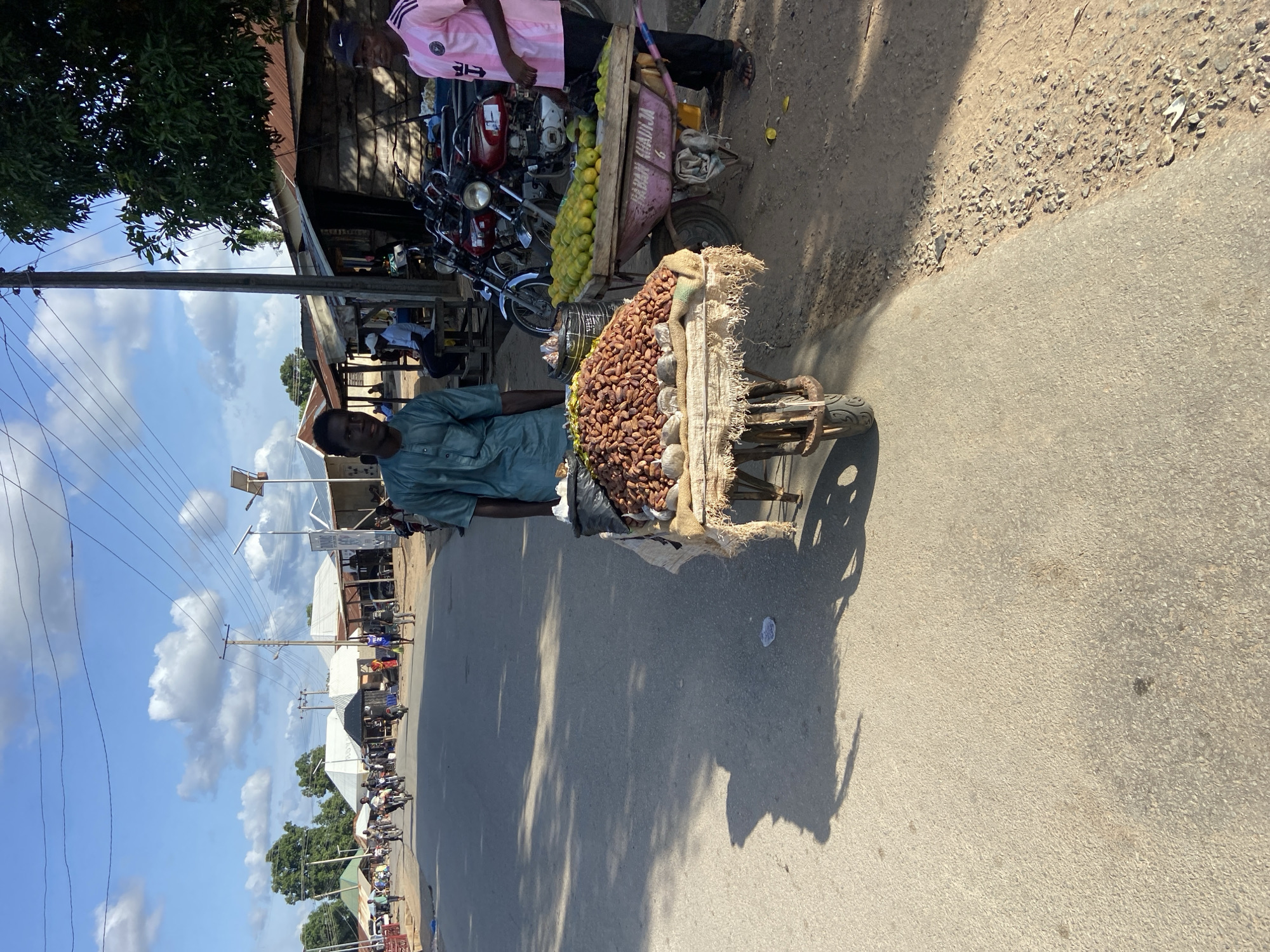 In Mararaba, Nigeria, a man stands beside a wheelbarrow filled with groundnuts. The wheelbarrow is parked on the side of a paved road, casting a shadow. Another wheelbarrow filled with fruits is nearby. The road is lined with shops and pedestrians. Overhead, power lines stretch across the scene. The sky is partially cloudy.