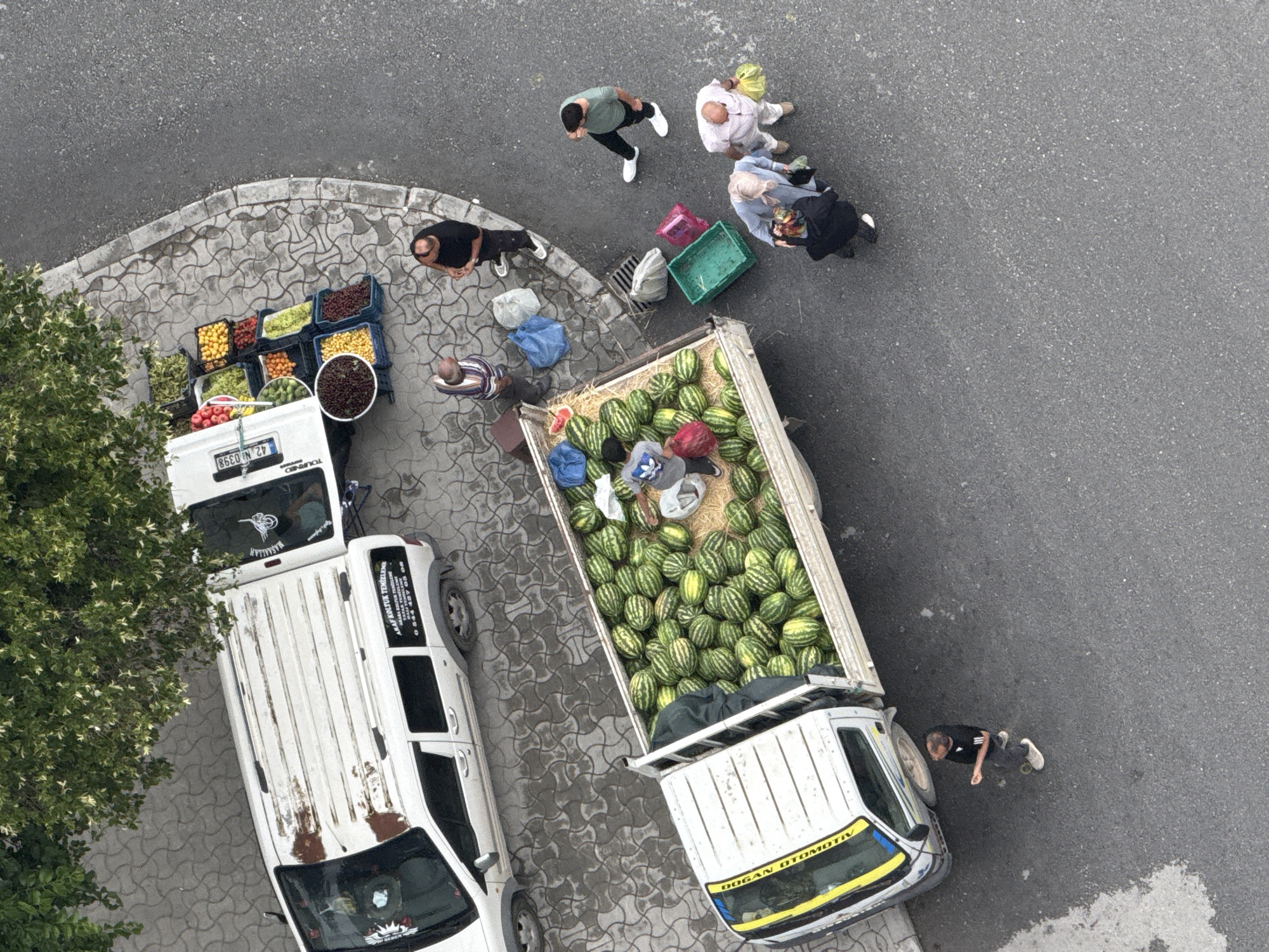 This aerial photograph, taken in Konya, Turkey, depicts a street scene with two vehicles, a produce stall, and several people. A white panel van is parked on the left, with its roof showing signs of rust and wear, and a sticker on the driver's side door displaying text. Adjacent to this van, a smaller white truck is positioned, its bed overflowing with watermelons. The truck also carries crates of assorted fruits, including grapes, tomatoes, oranges, and what appear to be lemons or limes, displayed on the side of the vehicle.
Several individuals are engaged in activities around the produce stall and the watermelon truck. Two men are standing near the front of the produce stall, seemingly interacting with each other. Further down the street, closer to the watermelon truck, a group of people are gathered. One person, wearing a gray hoodie with an Adidas logo, is handling a red object near the watermelons. Another person, in a black outfit, is squatting. Two other individuals are standing near a green crate, and another person is holding a yellow bag.
The ground is paved with patterned tiles near the produce stall and transitions to asphalt. The scene is shaded by the branches of a leafy green tree positioned in the upper left corner of the frame.
