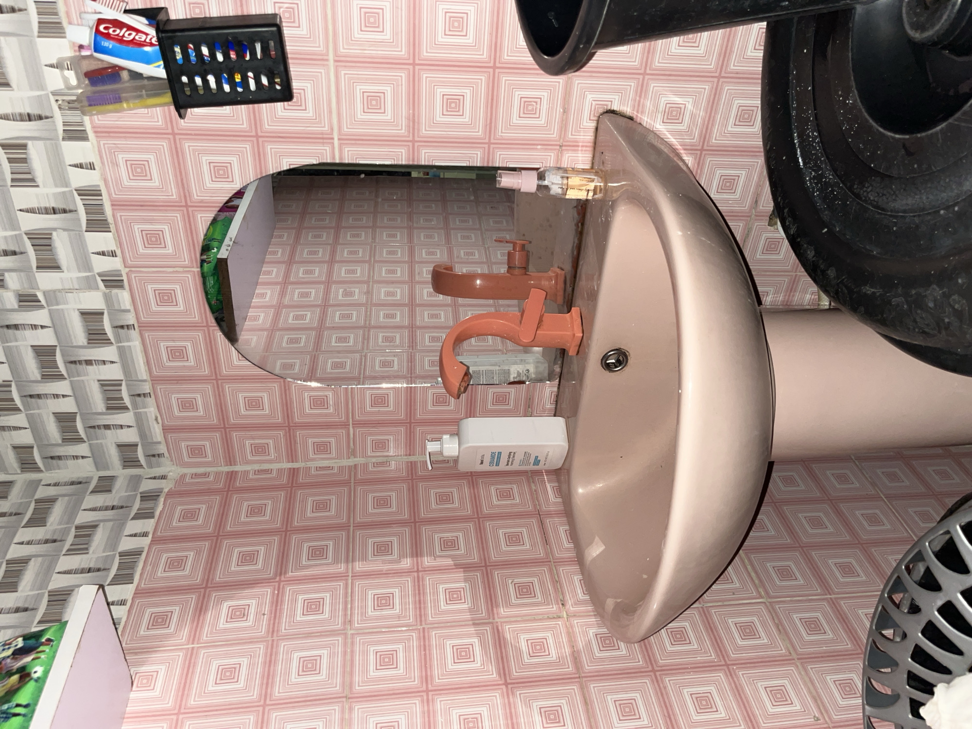 The image depicts a bathroom sink area in Abuja, Nigeria. The walls are covered in pink and white patterned tiles. A pink pedestal sink with a peach-colored faucet is the central focus. To the left of the sink is a white pump bottle of "Ceramide Hydrating Cleanser". To the right, a small clear bottle with a pink cap containing a light yellow liquid sits on the sink's edge. In the reflection of the mirror above the sink, another peach-colored faucet is visible. A black toothbrush holder containing toothbrushes and toothpaste is mounted on the wall to the upper right. Below the sink and to the right, a portion of a black trash bin is visible. In the lower left of the image, a silver mesh basket is partially visible.