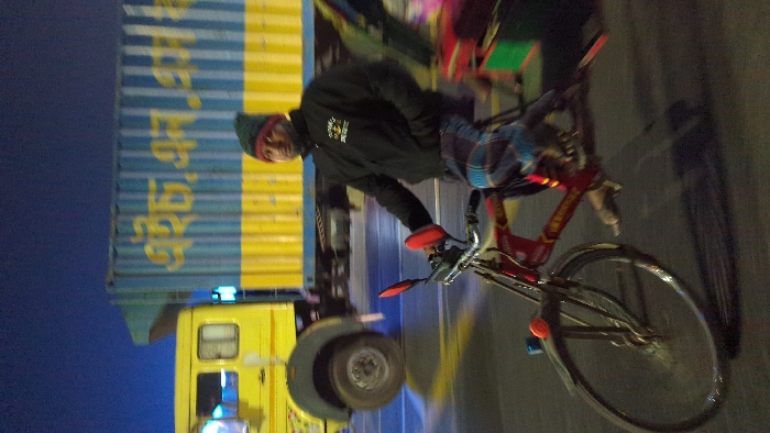 A man in a dark jacket and beanie rides a red bicycle down a road at night. A large yellow truck with blue writing on its side is parked in the background, facing the same direction as the cyclist. The road appears to be paved and illuminated by streetlights or vehicle headlights. The cyclist grips the handlebars with red gloves. The background is somewhat blurred, suggesting the cyclist is in motion.