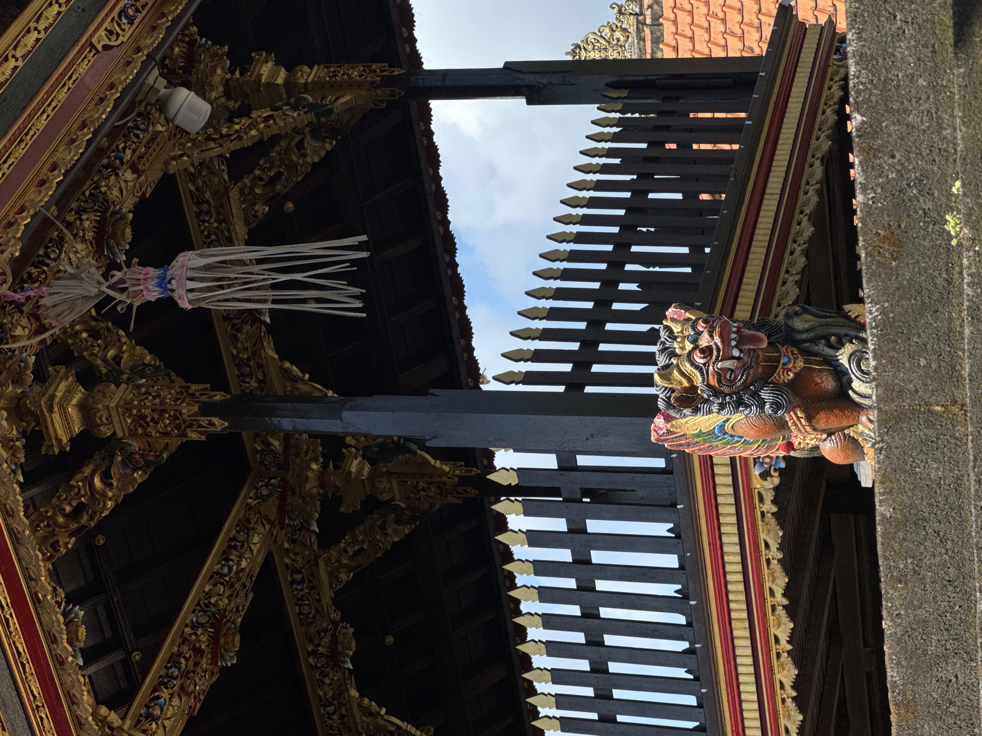 This is a low-angle shot of ornate architectural elements, presumably of a Balinese temple. The location is likely North Kuta, Indonesia, given the style of the architecture. The foreground features dark wooden beams and decorative carvings, painted in gold, red, and other bright colors. On the right side, there is a carved mythical figure, possibly a guardian statue, attached to a pillar. Through gaps in the wooden structure, patches of blue sky with some clouds are visible. A traditional Balinese offering, possibly a gebogan, hangs from one of the beams on the left. The overall scene is a close-up, detail-oriented view of Balinese craftsmanship and religious architecture.
