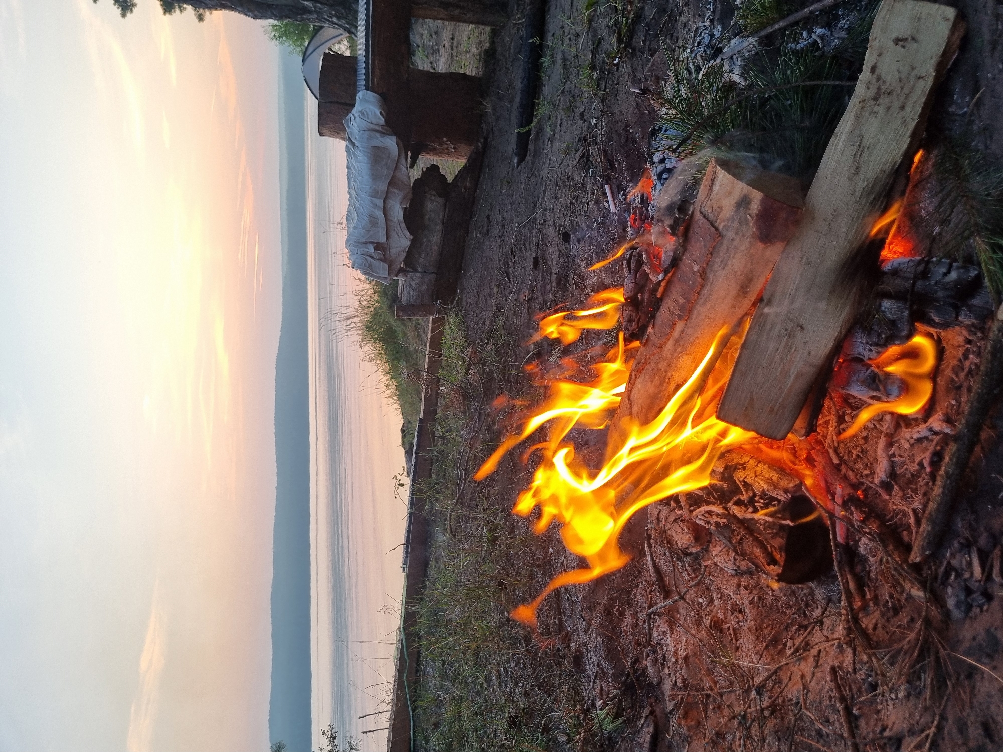 A campfire is burning on the shore of the Kama River in Chaykovsky, Russia. The sun is setting, casting a warm glow over the water and sky. In the foreground, the fire is crackling and producing flames that illuminate the surrounding logs and embers. The background shows a calm river with a distant tree-lined shore. A rustic wooden bench is visible to the right, covered with a light-colored cloth, and a tent is partially visible behind it. The scene conveys a peaceful, natural setting.