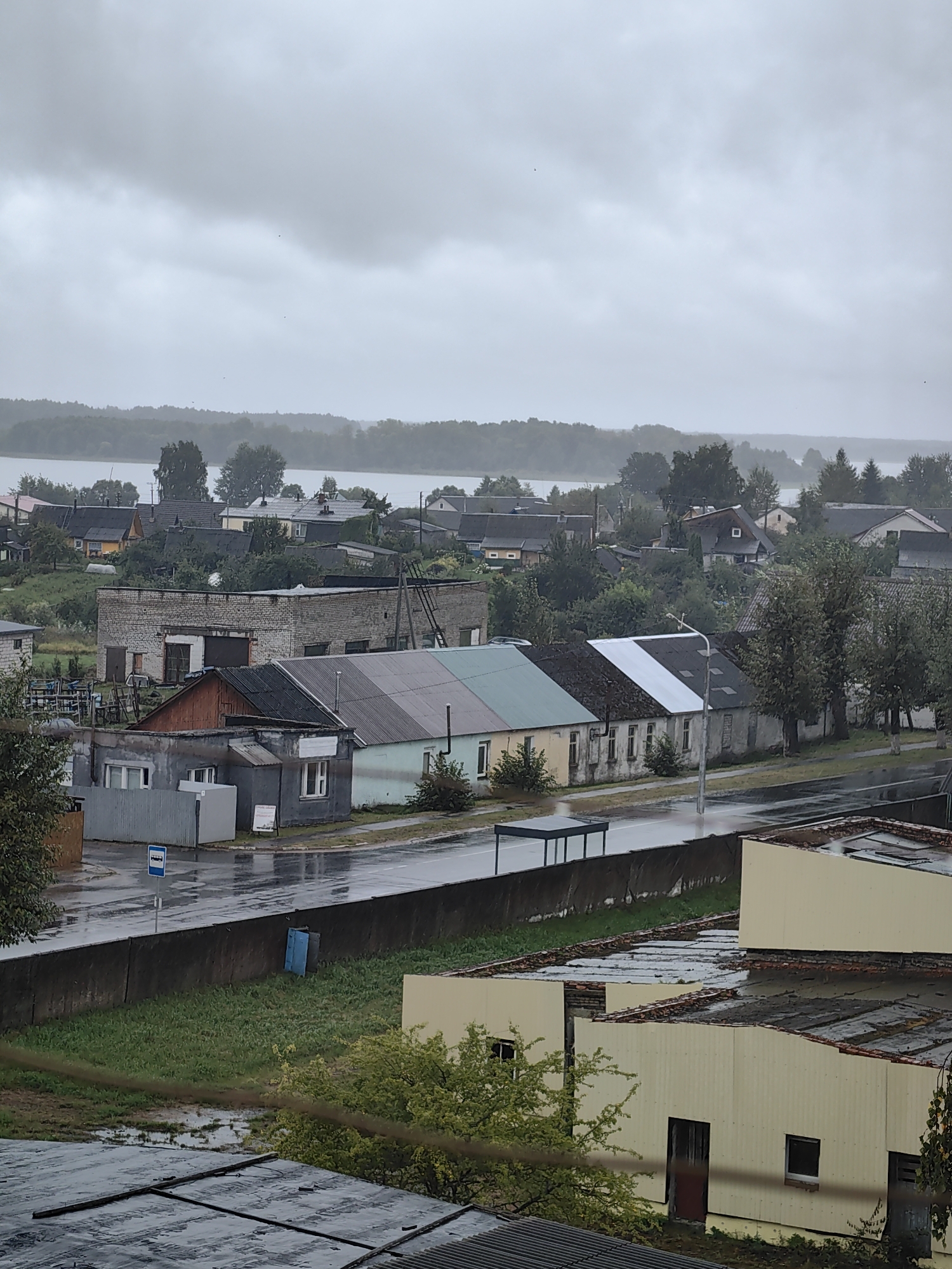 The image depicts a cloudy, overcast day in Myadzyel, Belarus, with a view overlooking a residential area near a body of water. The foreground features the dilapidated roofs of several industrial or warehouse-like buildings, constructed with corrugated metal and showing signs of disrepair. Beyond these structures, a street runs through the scene, wet from rain, with a blue bus stop sign and a covered bus shelter visible. A low, dark wall separates the street from a grassy area with puddles and a single blue dumpster. Further back, a row of low-rise houses with varied rooflines and muted colors lines the street. The houses appear to be a mix of single and multi-story structures, some with visible chimneys and dormer windows. Behind the houses, dense green trees form a treeline, interspersed with more buildings. In the distance, a wide expanse of water, likely Lake Myadel, stretches across the horizon, bordered by a further dense line of trees under a heavy, gray sky. There are no people visible in the image. The overall impression is one of a quiet, possibly rural, town on a wet and gloomy day.