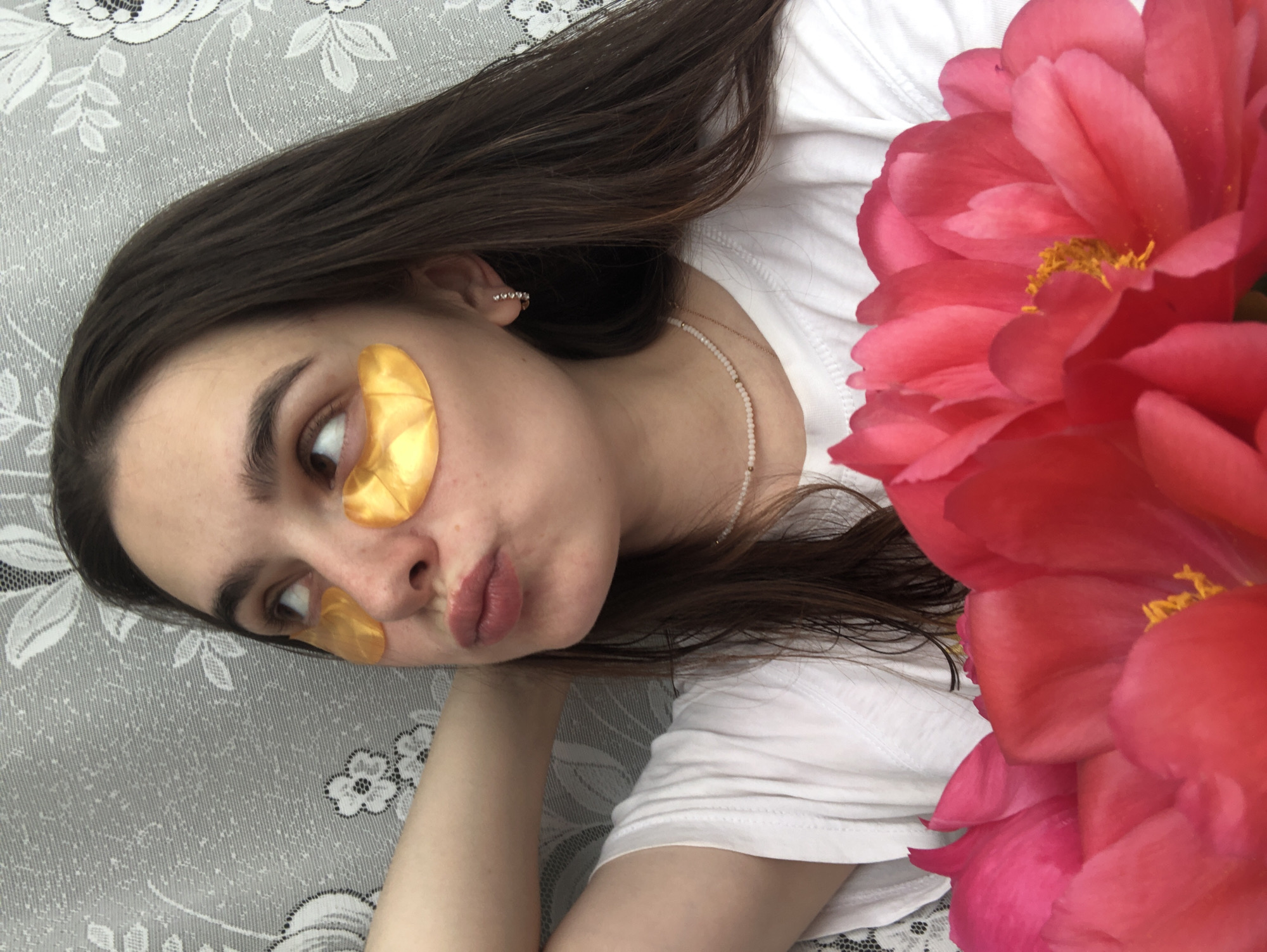 The image shows a close-up of a young Caucasian woman with long, dark brown hair. She is wearing a white t-shirt and has gold under-eye patches on. Her head is tilted upwards and to the left, with her right hand resting behind her head. She has her lips puckered as if she is about to kiss or is making a kissing face. In the foreground, there are several bright pink and red peony flowers. The background is a gray textured wallpaper with a white floral pattern. The lighting suggests an indoor setting. There is no information available to determine the specific location within Noviy, Russia.