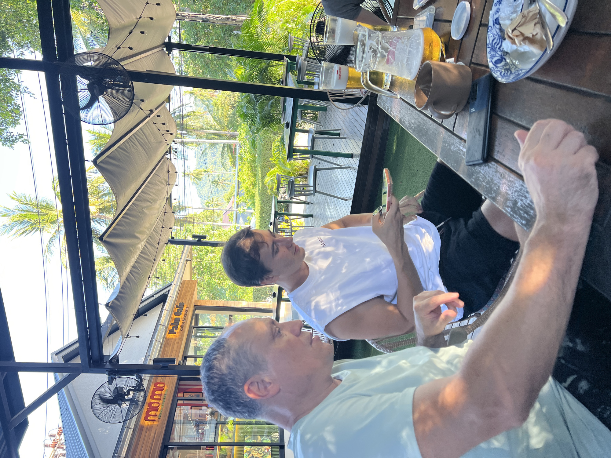 Two men are seated at a wooden table outside a cafe in Ban Bang Rak, Thailand, with palm trees and lush greenery visible in the background under a bright sky. The man on the left, wearing a light green t-shirt, is looking down at his phone. The man on the right, in a white t-shirt, is also looking at his phone, holding it in his hands. On the table are two glasses of beer, one with a pitcher, a brown mug, a mobile phone, and a plate with remnants of food and cutlery. In the background, fans are mounted on a structure with fabric awnings overhead. The cafe appears to be named "Mio Mio coffee & restaurants" and "Palm Cafe coffee & desserts".