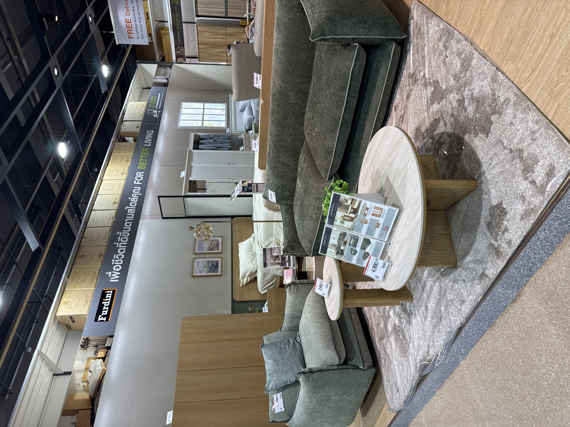 This image shows an indoor furniture showroom, likely in Baan Chaweng Noi, Thailand, featuring a staged living room area. The scene is illuminated by overhead lighting. In the foreground, a plush, dark green sofa is centrally placed on a light-colored, fluffy rug. To the left of the sofa is a round, marble-topped coffee table with a wooden base. Adjacent to the coffee table and to the left of the sofa is a matching armchair. Display stands with product information are placed near the coffee table.
Behind the seating area, a bed with white linens is set up, suggesting bedroom furniture is also on display. Further back, a large wardrobe with sliding doors is visible, as is a window with white panes. The showroom features a high ceiling with exposed ductwork and lighting fixtures. Various other furniture pieces and displays are visible in the background, including a tall floor lamp with a spherical shade and wooden cabinets. Signs for "Furdini" and "FOR BETTER LIVING" are prominently displayed on the wall behind the furniture, along with what appear to be product boxes stacked on shelves. A "FREE Delivery" banner is also visible in the upper right portion of the image. There are no people in the scene.