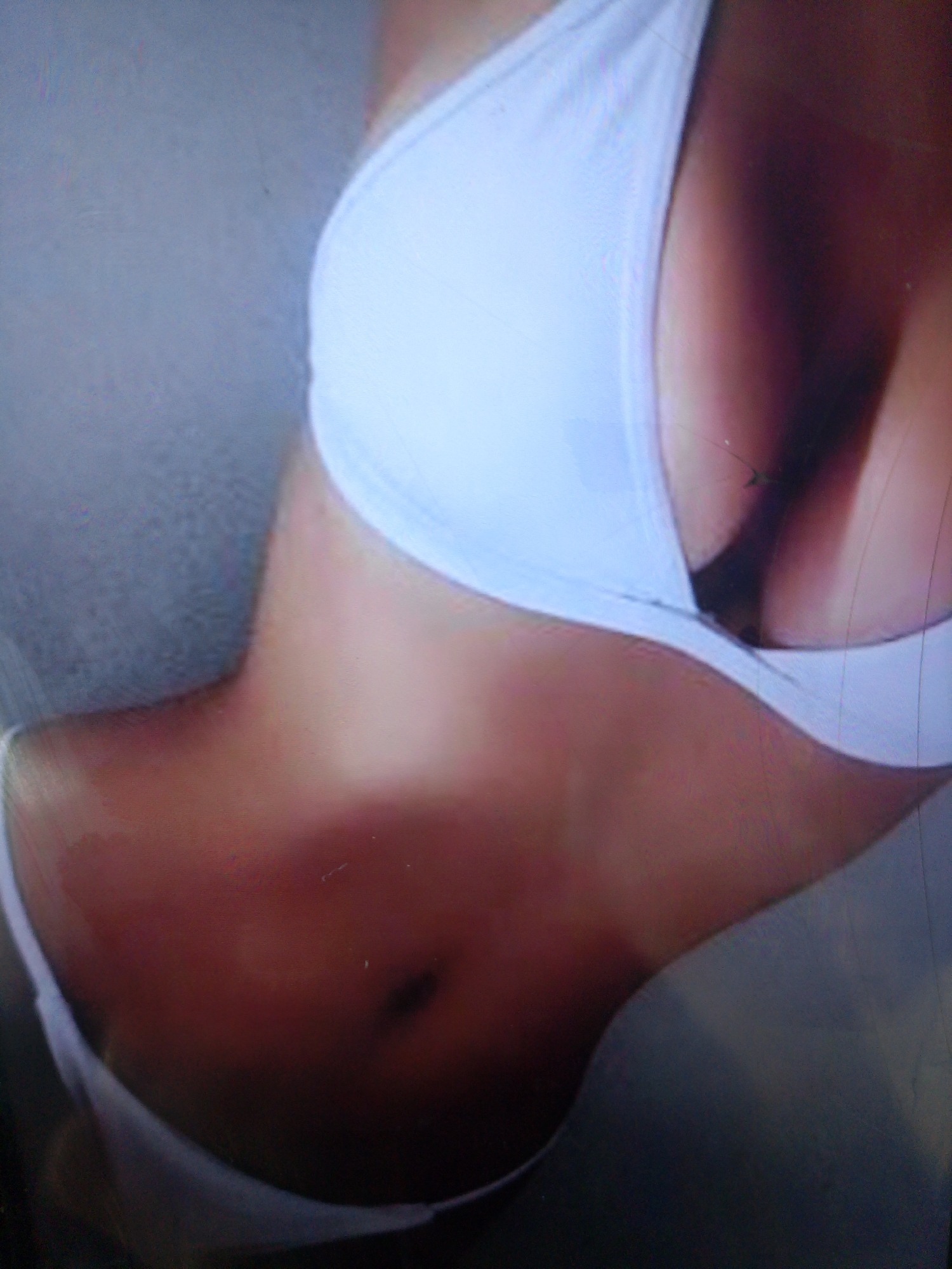 The image is a close-up, slightly blurry photograph of a person's torso, wearing a white bra. The person's skin tone is light to medium, and there are some shadows visible on the skin, suggesting a light source from the side. The background is a muted, gray texture. No specific location in Dobra Krynytsia, Ukraine, can be identified from this image.