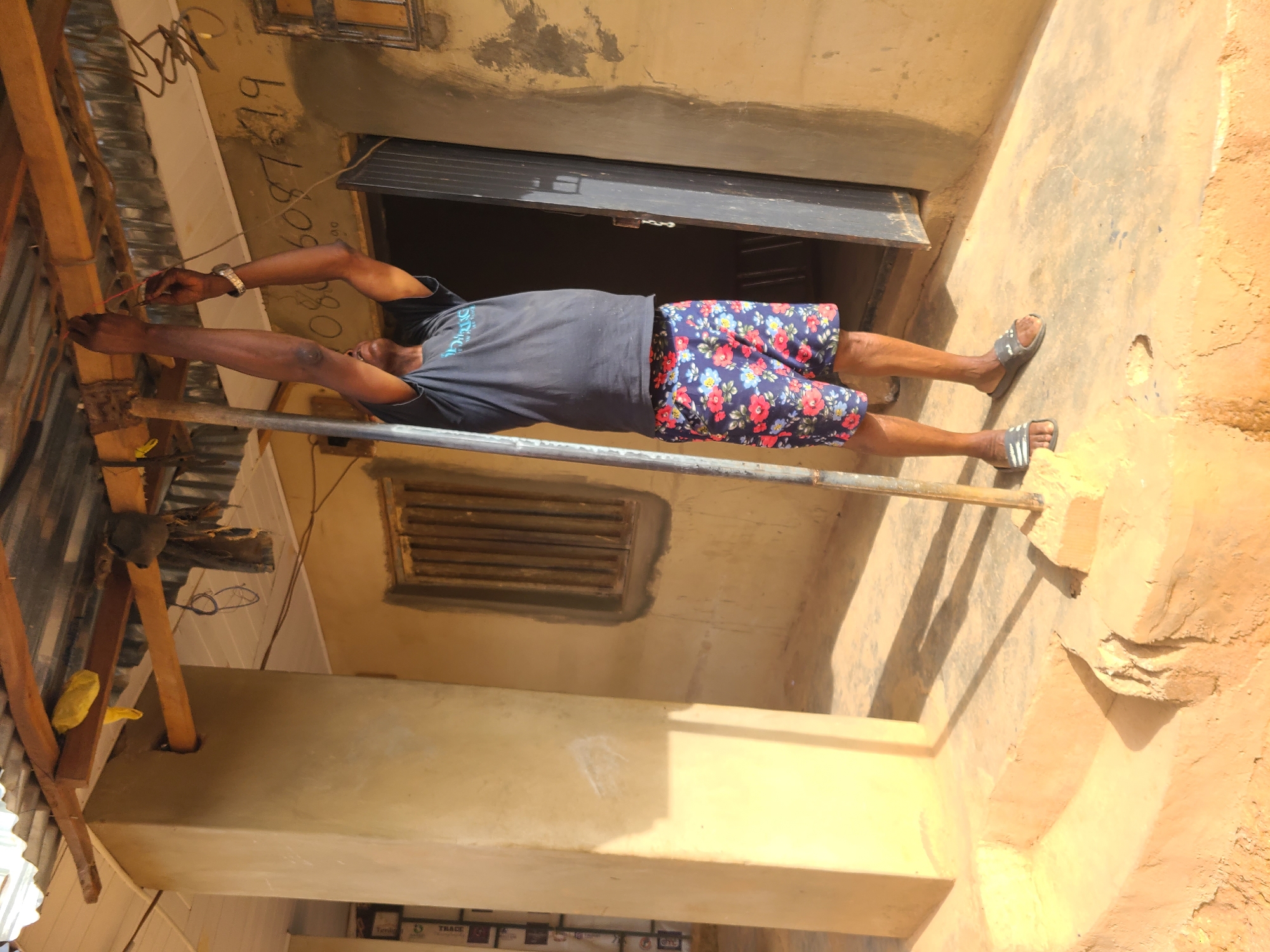 A man is standing on a metal scaffolding pole in Jos, Nigeria. He is wearing a dark gray t-shirt and colorful floral shorts and sandals. He is reaching up and working with electrical wires attached to a corrugated metal roof. There is an open window behind him and a louvered vent below. The building is tan with some exposed brick. There is text written on the wall above the window.