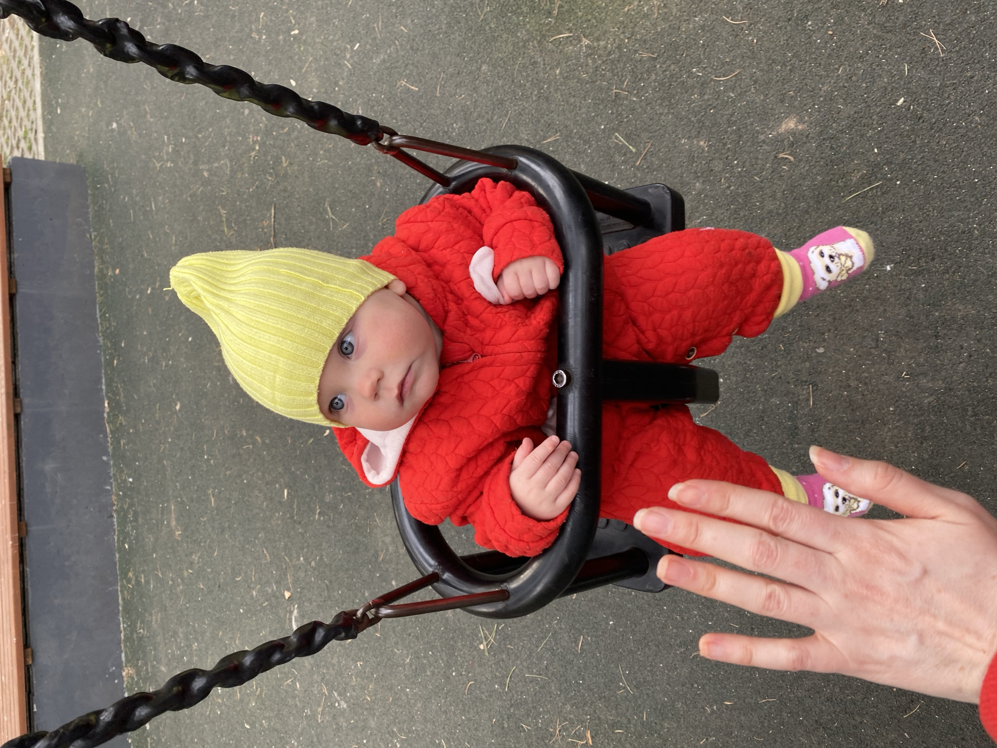 A baby sits in a black bucket swing at a playground, looking directly at the camera with wide, light blue eyes. The baby wears a bright red quilted snowsuit or bunting, a light yellow knitted beanie, and pink socks with yellow cuffs featuring a cartoon dog or bear character. The baby's small hands are visible within the suit.
An adult's fair-skinned hand is positioned on the right side of the frame, near the baby's foot, possibly preparing to push the swing or interact with the child. The swing itself is made of black plastic and metal, with sturdy chains extending upwards, partially visible.
The setting appears to be an outdoor playground, with the ground covered in a dark grey or green speckled rubberized safety surface, typical for such areas. Some dry grass or small debris is scattered on the ground. The lighting suggests an overcast day, as there are no harsh shadows. The baby's warm clothing indicates cool or cold weather. The overall impression is one of a caring interaction between the adult and baby in a safe play environment. The location context for this image is Yekekaterinburg, Russia. There is no discernible text visible in the image.