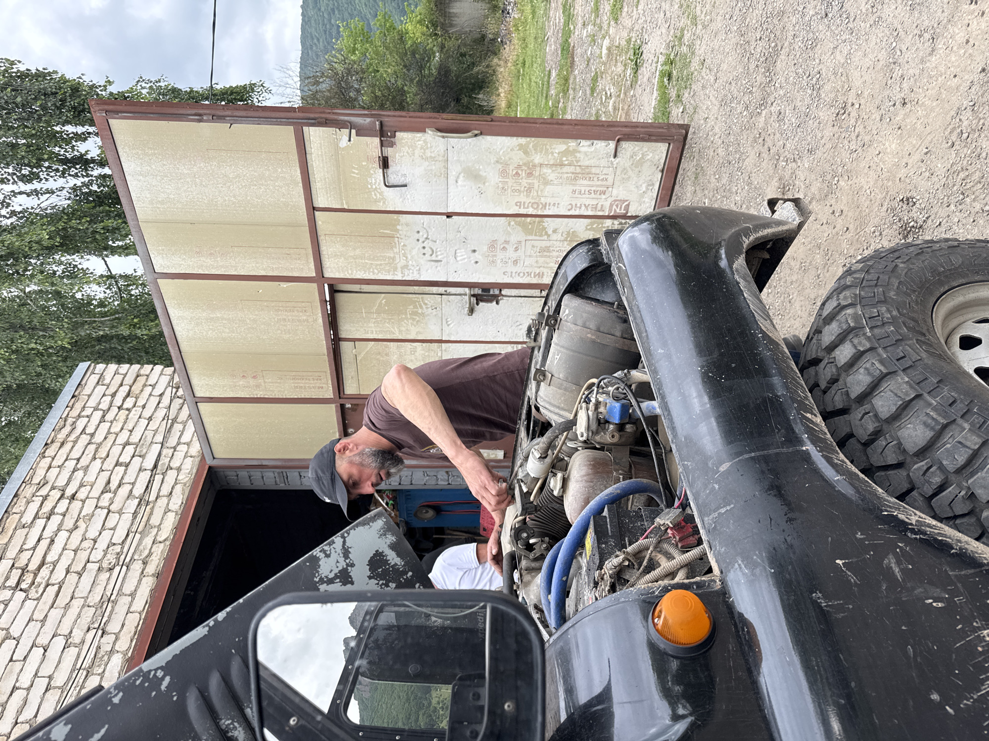 A man wearing a dark brown t-shirt and a black baseball cap is working on the engine of a black off-road vehicle. The vehicle's hood is open, revealing the engine components. Another person, wearing a white t-shirt, is partially visible in the background, seated inside the vehicle. The scene appears to be in an outdoor workshop or garage area, with a large metal garage door in the background. The ground is covered in gravel. In the distance, there are hills covered in green trees and a cloudy sky. The image is taken from a low angle, focusing on the vehicle and the man working on it. The location is identified as Karachayevsk, Russia.
