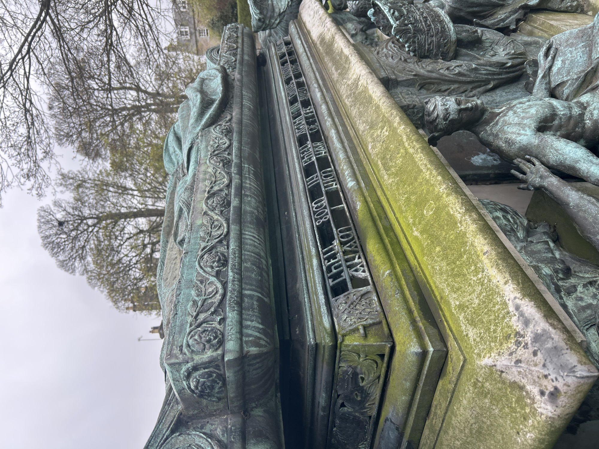 This image shows a section of a weathered, green-tinged bronze monument. The inscription "CHELL 8000 IN CHELL AGCH IN DHONS" is clearly visible, along with other indecipherable phrases due to wear and the angle of the photo. To the right of the inscription, there are sculptural representations of people. The overall environment suggests an outdoor location, likely within the city of Aberdeen City, United Kingdom.
