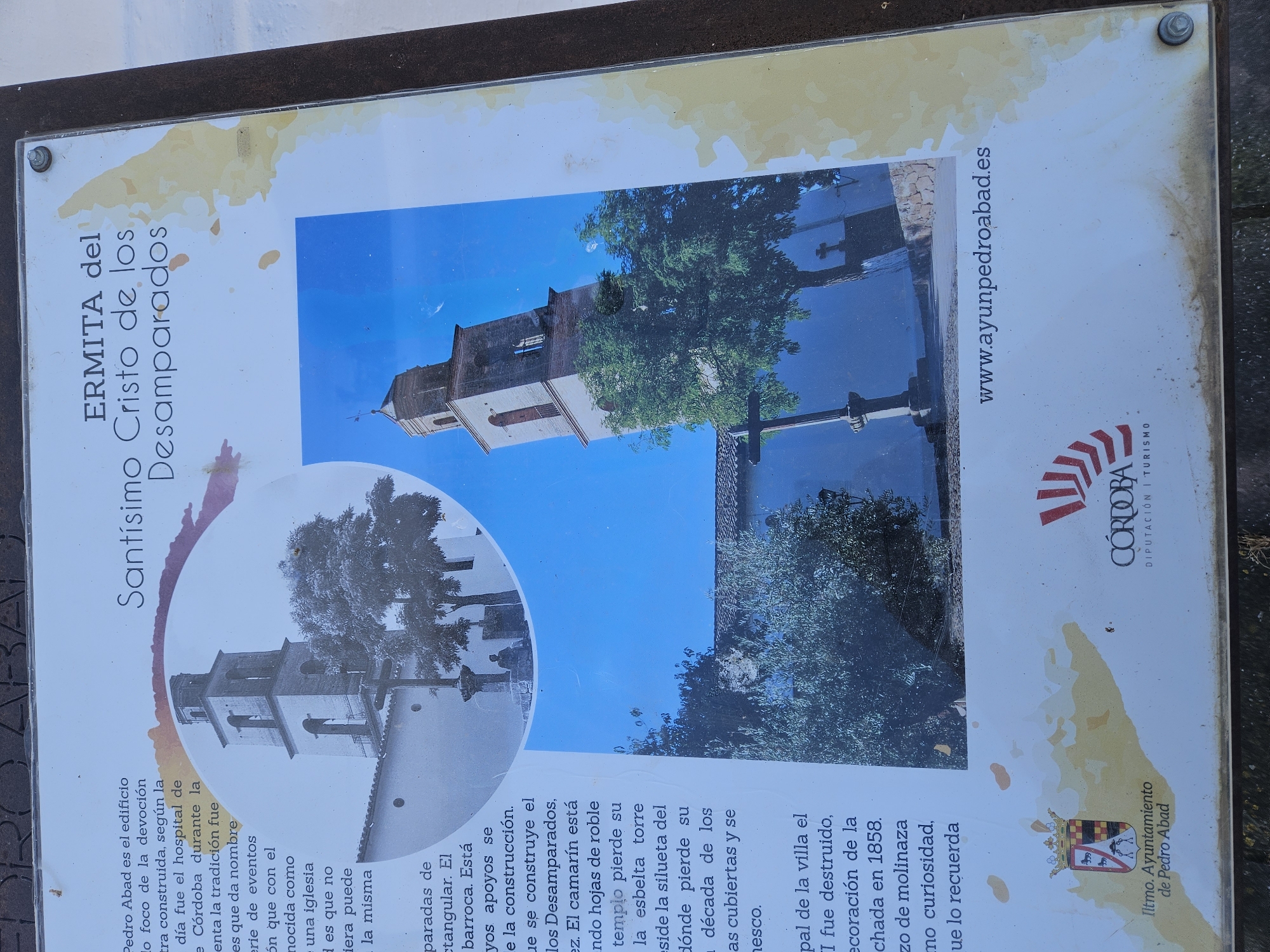 The image shows an information sign located in the city of Pedro Abad, Spain. The sign is about the "Ermita del Santísimo Cristo de los Desamparados" (Chapel of the Holy Christ of the Helpless). The sign features images of the chapel and text in Spanish detailing its history and significance. The sign also includes the website of the city council of Pedro Abad and the logo of the Córdoba tourism board.