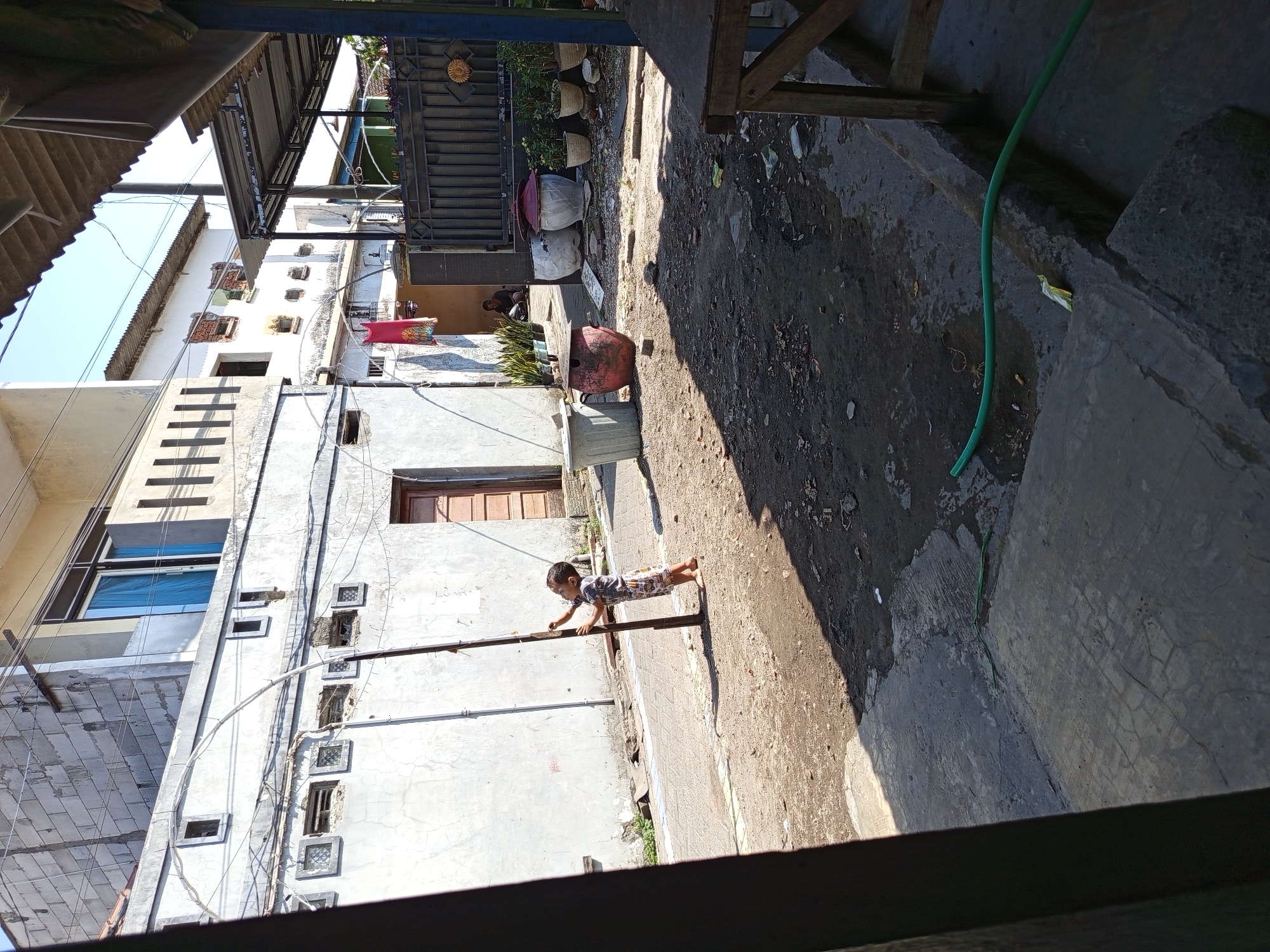 The image depicts a street scene in Tebel, Indonesia. In the foreground, a young child is standing barefoot next to a dark vertical pole, their right hand reaching up towards it. The child is wearing a patterned t-shirt and shorts. The ground is composed of dirt and concrete, with a green hose snaking across the lower right portion of the frame. In the background, a white building with several small square windows is visible, along with a brown wooden door. Further back, another building with a corrugated roof and exposed brickwork can be seen. A dark metal gate is present on the right side of the image, and behind it, two large terracotta pots are situated. A person is seated inside a doorway behind the gate. The environment appears to be a residential or commercial area, likely in a warm climate given the bright sunlight and sparse vegetation.