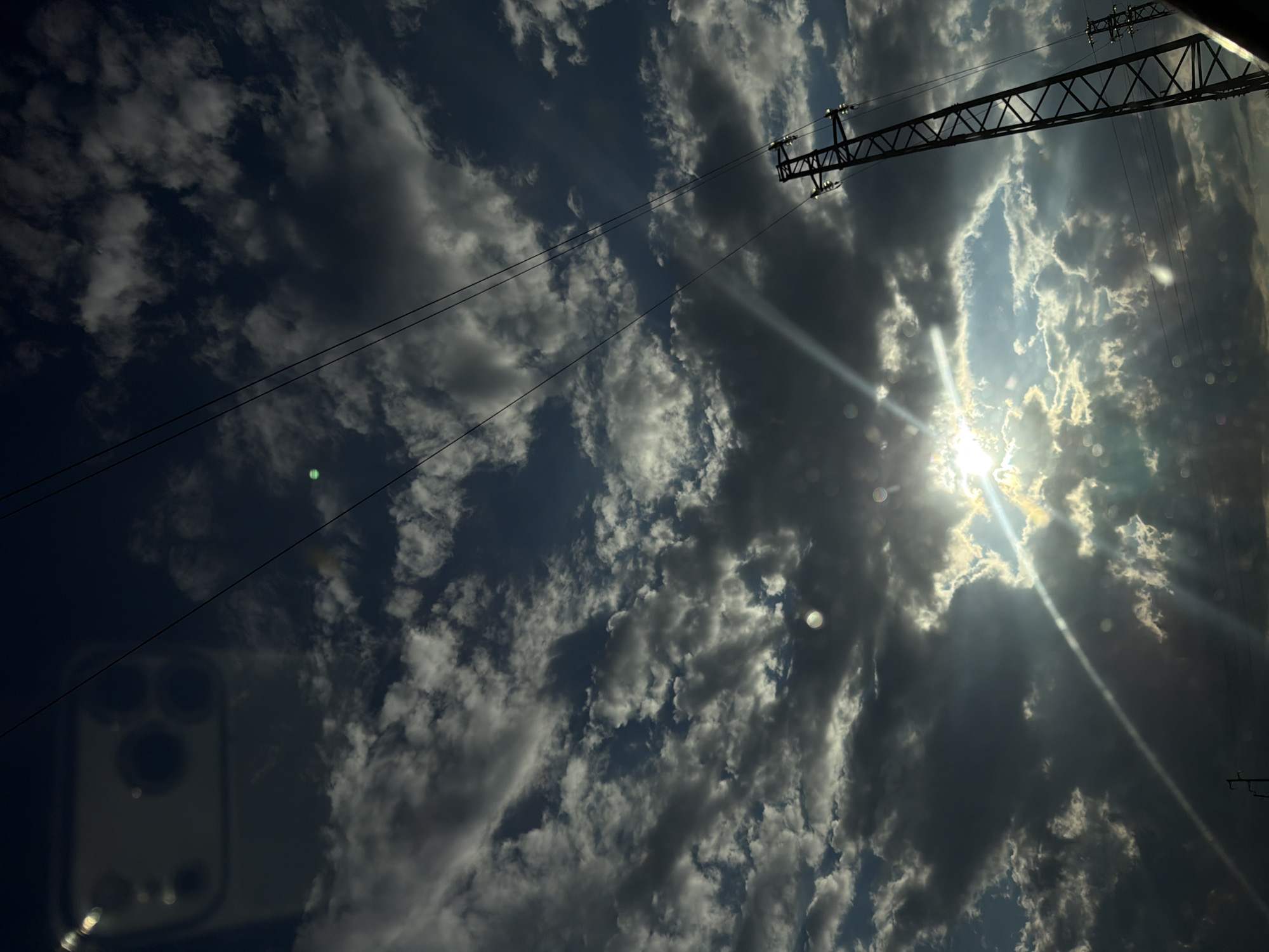The image depicts a sky view in Eleshnitsa, Bulgaria, dominated by an overcast yet dynamic cloud formation with areas of clear blue sky and direct sunlight. The sun, partially obscured by clouds, generates prominent lens flares and radiant sunbeams that penetrate the cloud cover, creating a dramatic interplay of light and shadow. Multiple circular lens artifacts are present across the frame, including a distinct bright green orb in the upper-mid left quadrant. In the upper-left, a faint, blurred reflection of a smartphone, identifiable by its multi-camera array, is visible, suggesting the photograph was taken through a reflective surface such as glass. Several dark, parallel power lines traverse the sky from the upper-left to the mid-right. A large, dark-silhouetted metal lattice-structure power transmission tower stands prominently on the right side of the frame, extending from the middle to the bottom-right, with its insulators faintly illuminated. A segment of a second, similar tower is visible further to the right edge. No individuals or specific actions are discernible. The overall scene captures an atmospheric sky with industrial infrastructure.