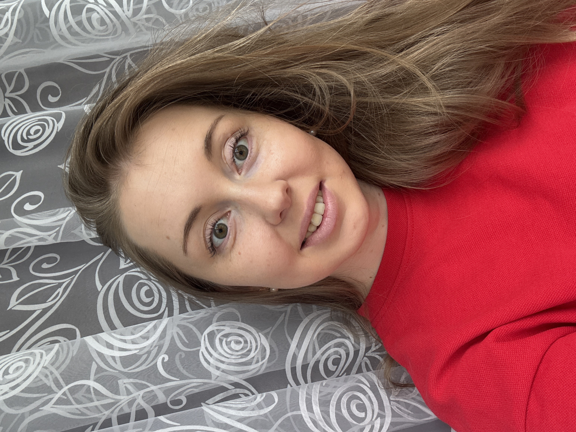 A woman with long brown hair and fair skin is lying on a surface with a gray and white patterned fabric. She is wearing a red sweater. The location is Unknown, Unknown.