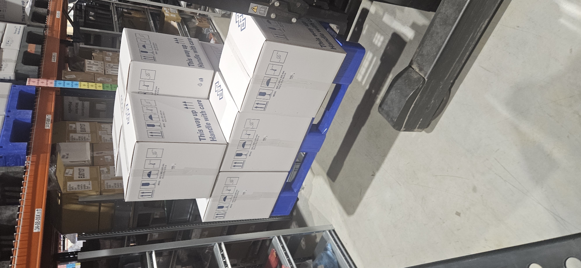 A stack of white cardboard boxes rests on a blue plastic pallet, captured from an overhead perspective within a warehouse. The tines of a forklift are prominently visible in the foreground, indicating that the pallet is either being moved or has just been positioned. The environment is an indoor storage facility with metal shelving units visible in the background, holding various other packed goods and boxes.
Each white box displays standard shipping and handling symbols, including upward-pointing arrows ("This way up"), an umbrella (for "keep dry"), a broken glass icon (for "fragile"), and symbols indicating stacking limitations. Accompanying text on the boxes reads "This way up" and "Handle with care." Dimensions are also printed, such as "MEAS: 405 x 385 x 425 mm." and "15.8 x 15 x 16.6 in."
Labels and text on the background shelving and other items include "34-08-09X1", "34-07-09XT", and multiple instances of "EU87137". Additional text like "Plovdiv", "Markov", and "Konstantin" can also be seen on some items. The presence of "Plovdiv" helps confirm the location in Plovdiv, Bulgaria. The lighting appears to be artificial, typical of an indoor industrial setting.