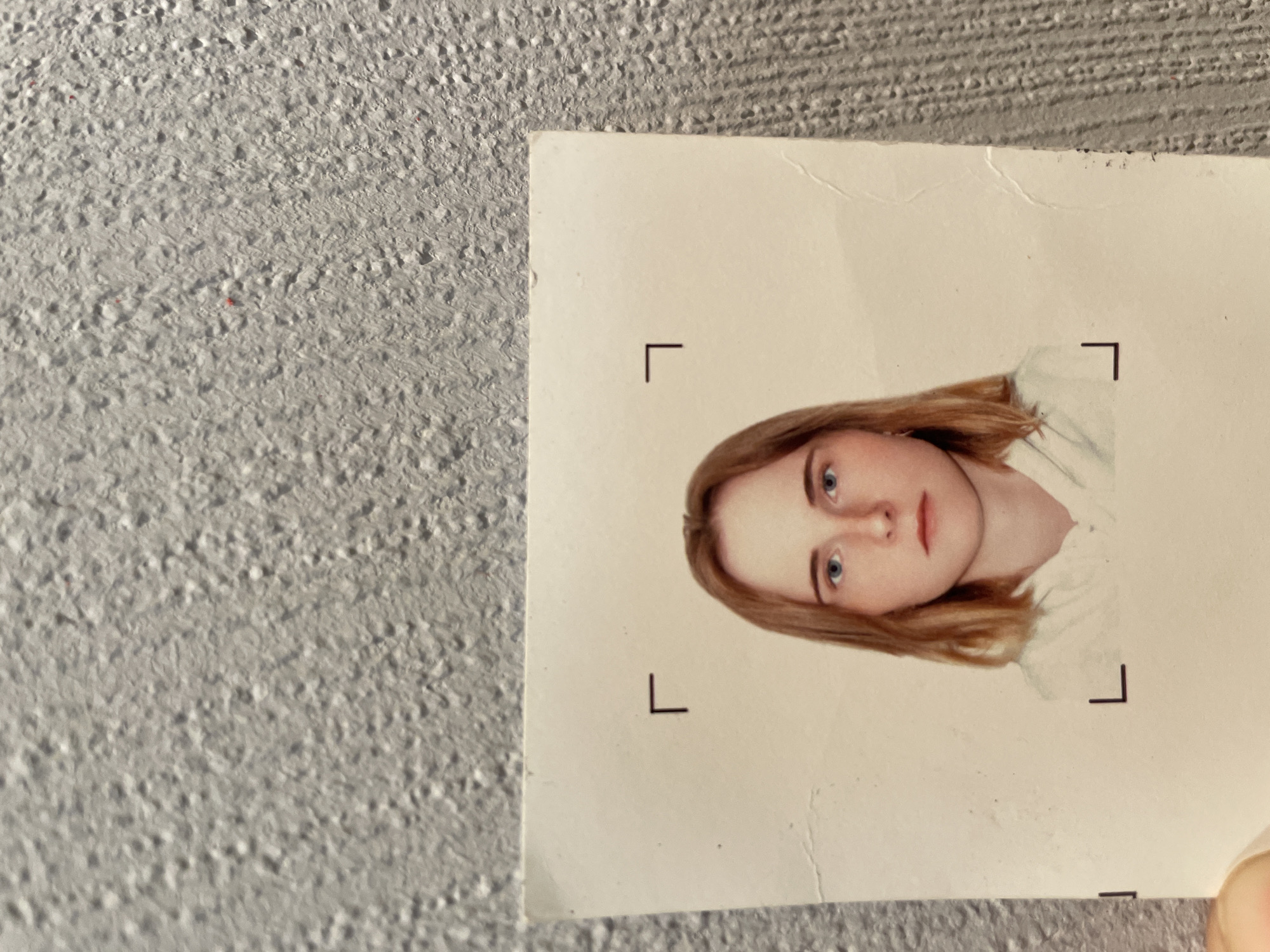A passport-style photograph of a young Caucasian female with shoulder-length light brown hair and blue eyes is shown. She is looking directly at the camera with a neutral expression. Her attire consists of a light-colored hooded sweatshirt. The photograph is printed on a light-colored paper with dark corner registration marks. The background behind the subject is a textured gray surface, likely a wall. The photograph appears to be part of a larger document or album.