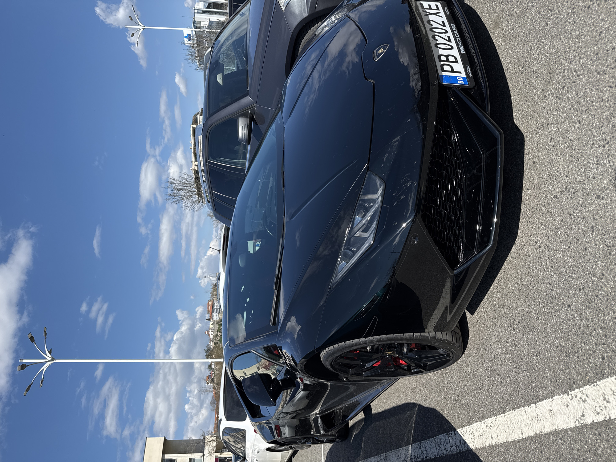 A black Lamborghini Huracan is parked in a lot next to a gray SUV. The Lamborghini has a Bulgarian license plate reading "PB 0202 XE" and red brake calipers. The parking lot is paved with asphalt and has white parking space lines. A tall white street lamp is visible in the background. The sky is blue with scattered white clouds. The photo appears to be taken in Plovdiv, Bulgaria.