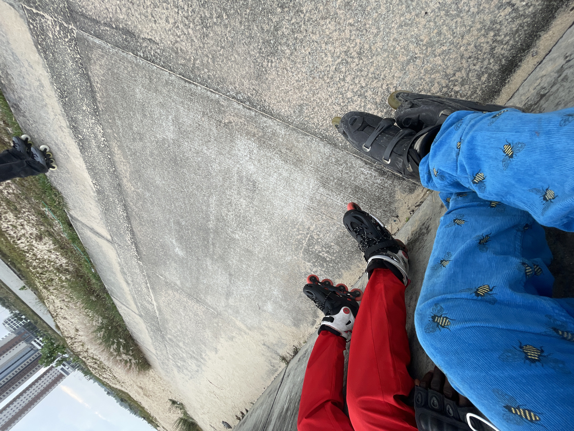 Two individuals are seated on a concrete embankment, wearing rollerblades. The person on the left wears red pants and black and white rollerblades with red wheels. The person on the right wears blue corduroy pants with a bee pattern and black rollerblades. Both individuals are facing away from the camera. In the background, there are buildings, grassy areas, and a body of water. The scene is located in Inupa Kekere, Nigeria.
