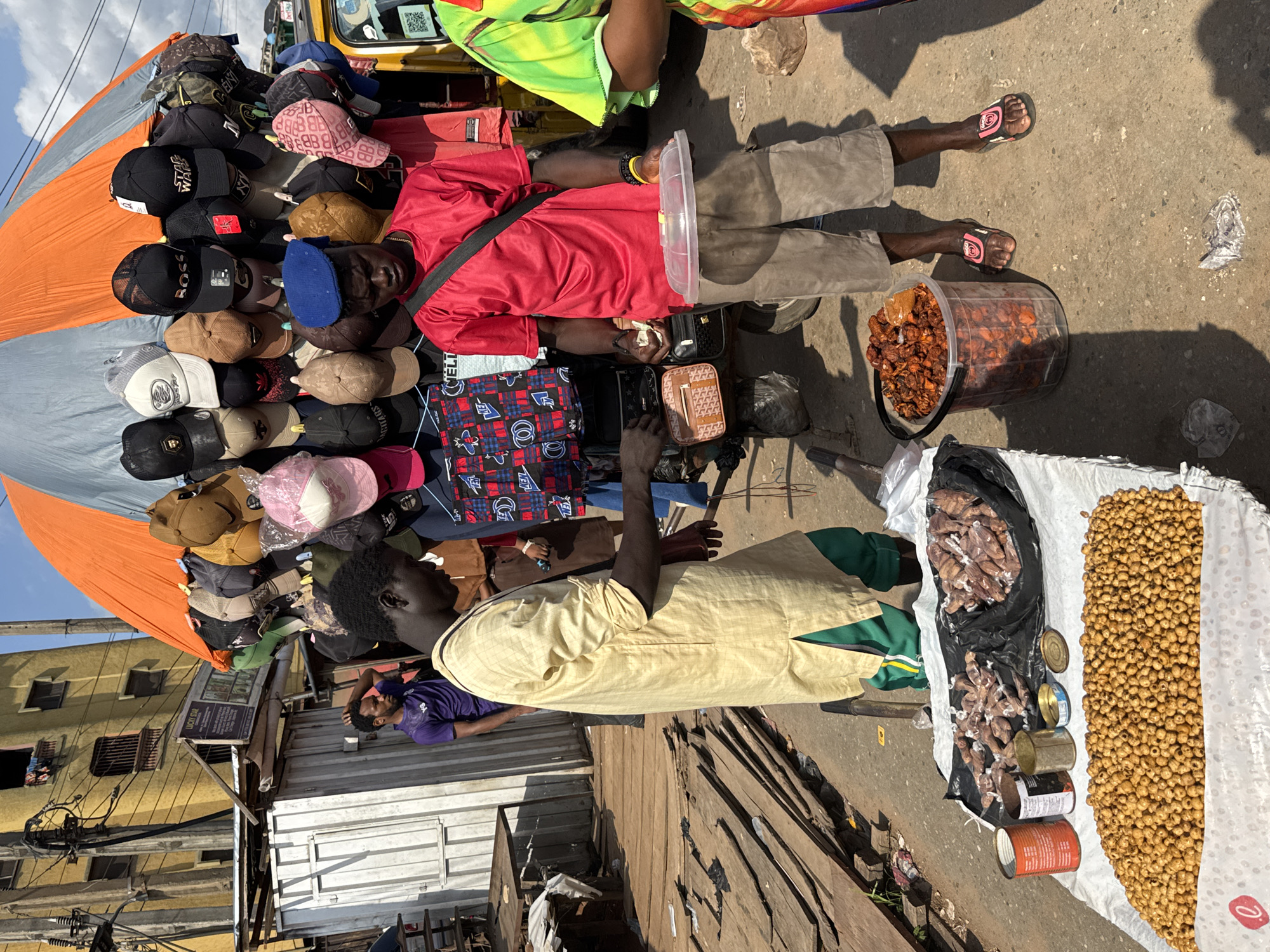 The scene depicts a marketplace in Nkpor, Nigeria. Two men are interacting at street vendor stalls. One man wears a red shirt and khaki shorts, a black strap across his chest, and slides on his feet; he stands next to a clear plastic bucket filled with a dark substance. The other man wears a light yellow long-sleeved shirt over a green garment; he stands beside a white bag filled with small, round yellow objects, several tin cans, and a black bag containing packaged items. Various hats are displayed under an orange and grey awning. A man in a purple shirt sits in the background. The stalls are set up on a wooden platform next to a paved road. A building is visible in the background.