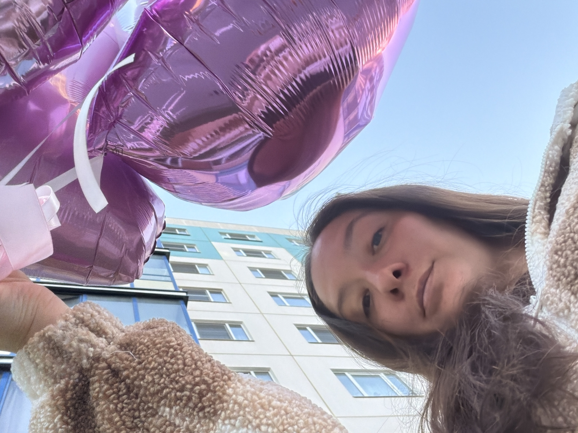 A low-angle selfie shows a young woman holding pink and purple heart-shaped balloons in front of a building in Tokkari, Russia. She wears a fuzzy brown and white jacket. The building behind her is multi-story with a white facade and rows of windows, some framed in blue. The sky is light blue.