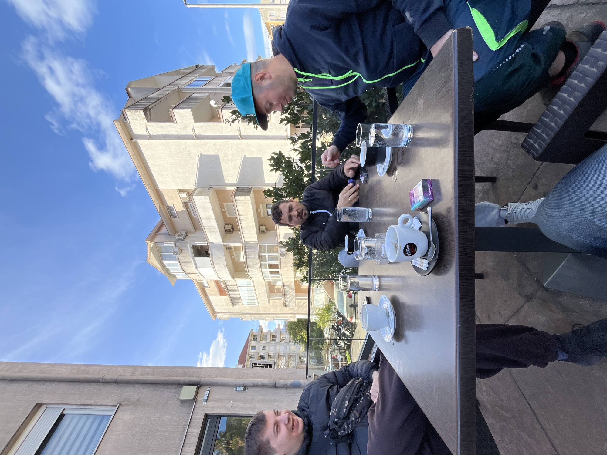 Four people are sitting at a brown table on a patio. Three empty coffee cups and three glasses of water are on the table, along with a pack of cigarettes. A man in a black jacket is seated on the lower left side of the frame, smiling. Directly across from him, a man in a black jacket is looking at a phone. To the right of him, a man in a blue cap and a blue and black jacket leans forward, looking at the pack of cigarettes. Another person is seated on the right side of the frame, with part of their legs and jeans visible. In the background, a multi-story building with balconies and air conditioning units is visible under a blue, cloudy sky. The scene appears to be in an urban setting, likely Budva, Montenegro, given the location information.
