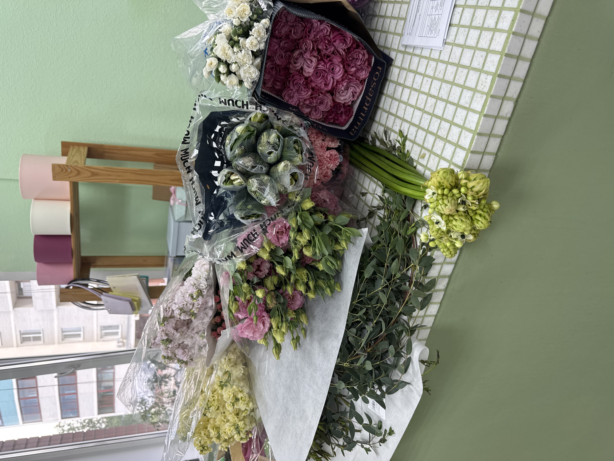 This image captures an indoor setting, likely a flower shop or a preparation area, filled with a diverse collection of fresh cut flowers. The scene is dominated by numerous bouquets and bundles of flowers, some wrapped in plastic and others in paper or specialized packaging, indicating they are either newly arrived stock or ready for arrangement.
The subjects include a variety of flowers: a bunch of white spray carnations or small roses, a box full of deep pink to maroon roses with the brand "rosaprima" visible, and bundles of light green, tightly budded flowers, possibly ranunculus or similar. Other visible flowers include pink carnations, clusters of pink and purple lisianthus with green buds, light pink stock flowers, and yellow stock flowers. Several bundles of fresh green foliage, possibly eucalyptus, are also present, some wrapped in white paper.
The flowers are arranged on what appears to be a white tiled surface with light green grout, contrasting with a solid light green painted wall. In the background to the left, a wooden shelf holds rolls of pink and white wrapping paper, suggesting materials for floristry. A window offers a view of urban buildings outside, featuring multiple stories and various window configurations, characteristic of a daytime cityscape in Izhevsk, Russia.
Visible text includes the brand "rosaprima" on the box of maroon roses and repeated text "MUCH MUCH MUCH" on a dark-colored plastic wrap. A document with printed text is also visible on the tiled surface, though its contents are unreadable. The overall impression is one of a busy and organized workspace dedicated to floristry.