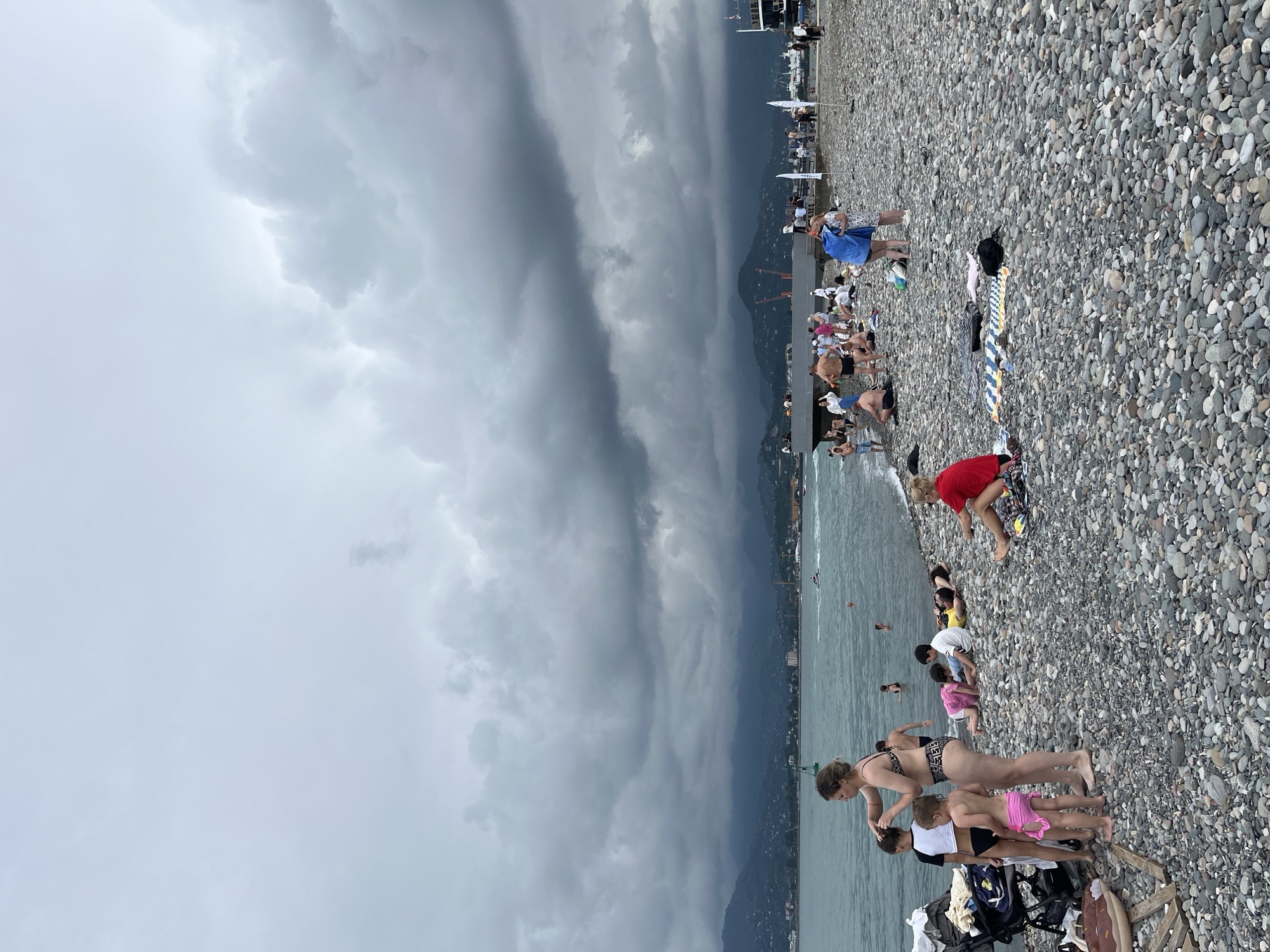 The image shows a crowded pebble beach on a cloudy day in Batumi, Georgia. In the foreground, several people are scattered across the beach, some sunbathing, others talking or interacting with children. A woman in a bikini is attending to two young girls, one in a white t-shirt and black bikini bottoms, the other in a pink bikini. A stroller is parked nearby. Further down the beach, more people are visible, some are in the water, while others are sitting on towels. In the background, a pier extends into the sea with more people and several boats. The sea is a deep blue-green, and beyond it, a city skyline with mountains in the distance is visible. The sky is heavily overcast with dark, dramatic clouds. The overall impression is a busy, albeit somewhat somber, beach scene.
