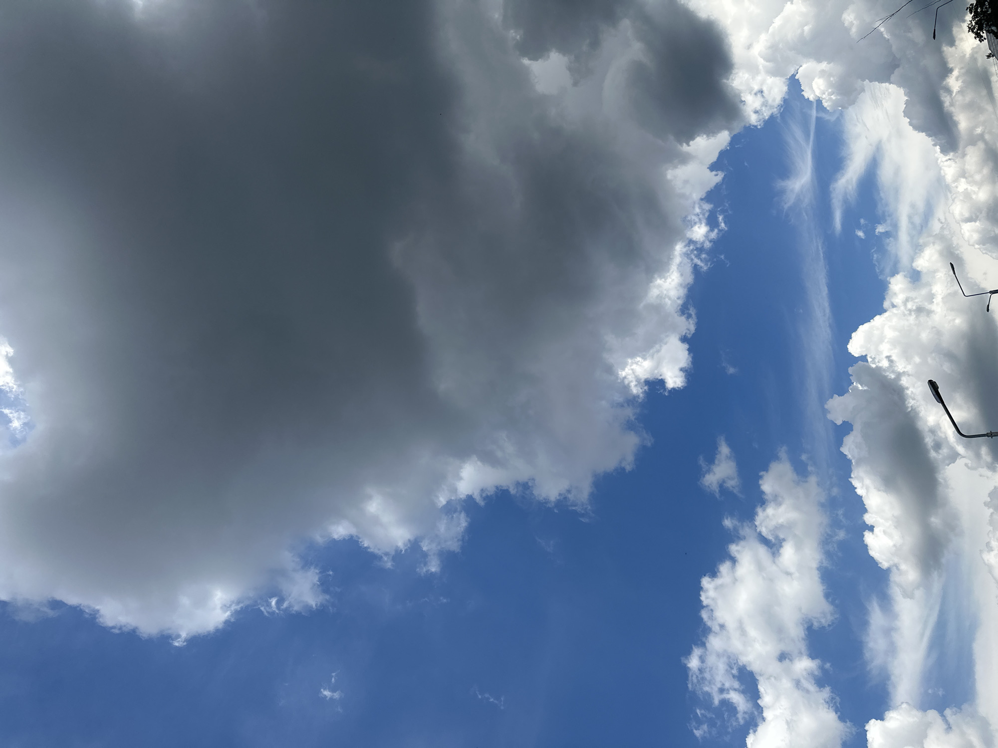 The media file captures a dynamic sky over Lopaskyne, Ukraine. The scene is dominated by a dramatic interplay of light and shadow created by various cloud formations. A large, dark gray cloud mass occupies the left side of the frame, suggesting impending rain or a storm. This is contrasted by bright, white cumulus clouds scattered across the blue sky, particularly on the right and towards the center. Wispy, cirrus-like clouds are also visible, adding texture and depth to the celestial panorama.
The orientation of the image, with the sky filling the entire frame, implies the shot was taken looking upwards from ground level. The presence of streetlights, one prominently visible on the right side of the image, along with what appears to be the top of a tree or building on the far right, provides subtle location cues, suggesting an urban or semi-urban setting.
The time of day is likely daytime, given the brightness of the sky and the illumination of the clouds. The weather appears to be mixed, with both stormy and fair-weather clouds present. This could indicate a transitional period, perhaps before or after a rain shower. No people are visible in the media file. The overall mood conveyed is one of natural grandeur and the ever-changing beauty of the sky. No text is visible in the image.
