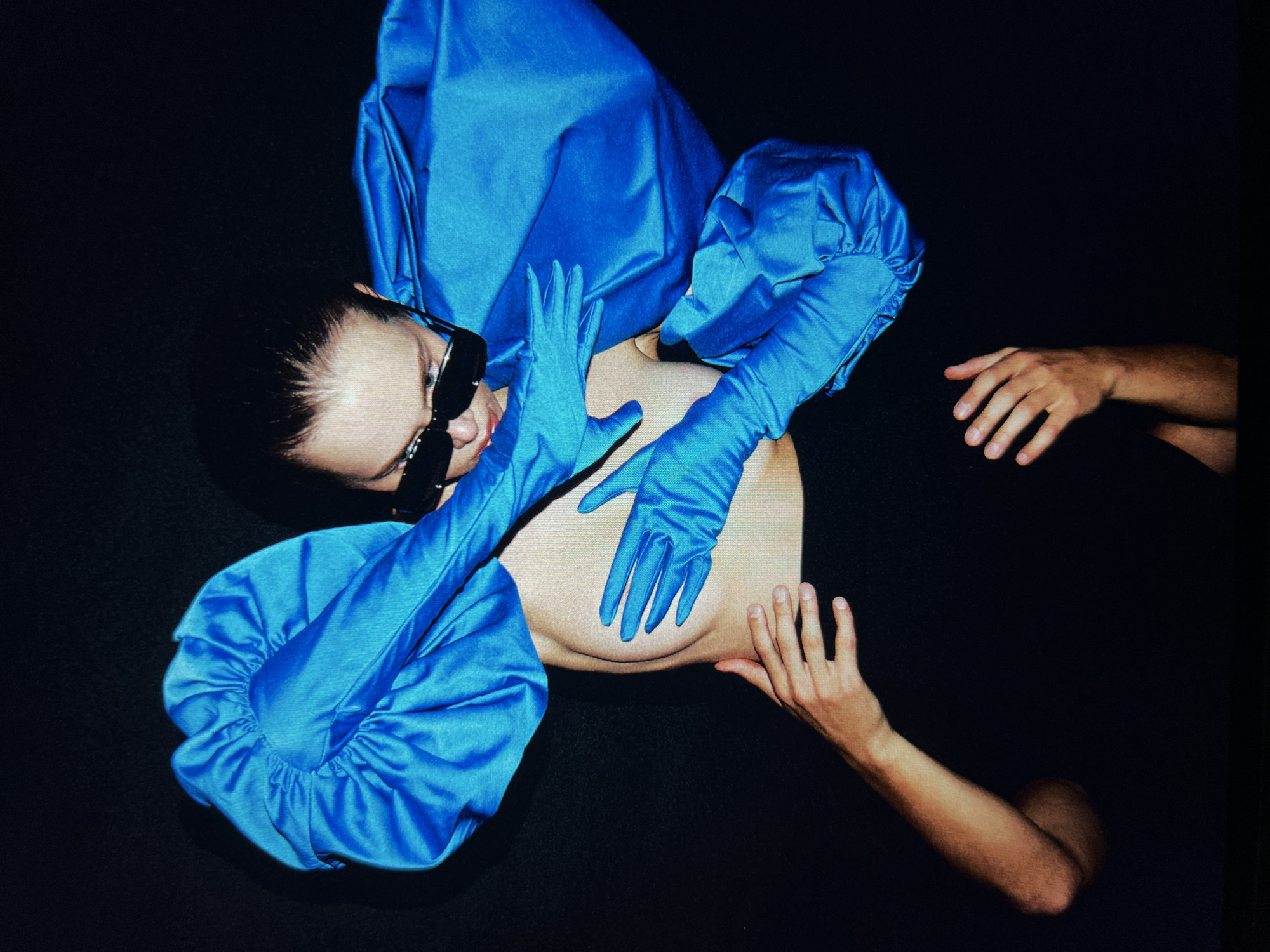 A high-angle shot shows a person wearing a black top, black sunglasses, and voluminous bright blue sleeves and gloves, against a black background. Their right hand, clad in a blue glove, is placed over their chest, while their left arm, also in a blue glove, curves around their head. Another person's hands are visible at the bottom, with their right hand resting on the upper thigh of the person in front and their left hand open.