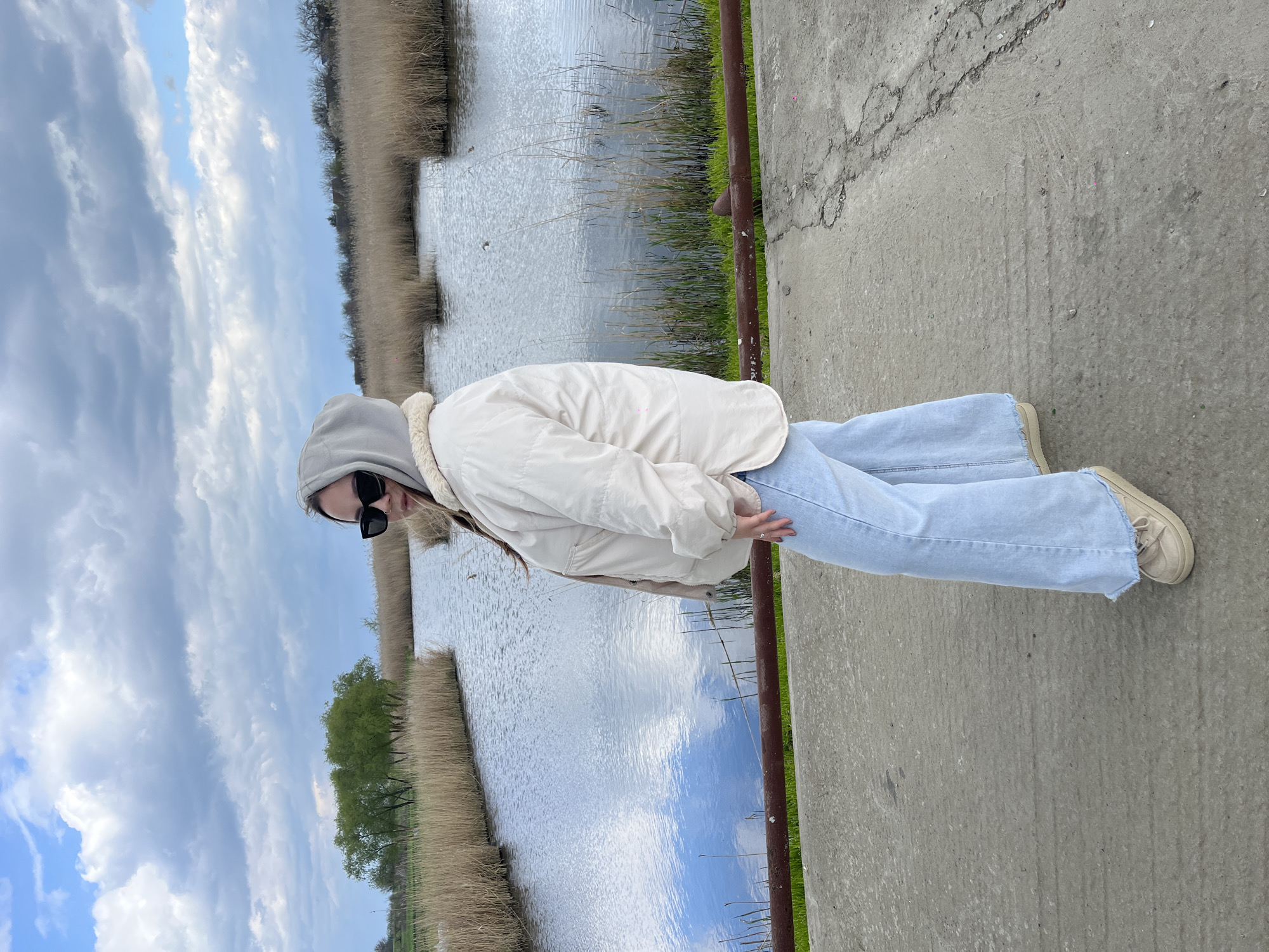 A young woman stands outdoors on a concrete path near a body of water. She wears a light-colored hooded sweatshirt, a light beige jacket, light-blue jeans, and beige sneakers. She also wears sunglasses. The location appears to be near a lake or river with reeds and vegetation on the banks. The sky is cloudy with some blue patches. The concrete path has a metal railing with grass growing at the base.
I am unable to provide the specific location as Novopokrovskaya, Russia, from the visual information.