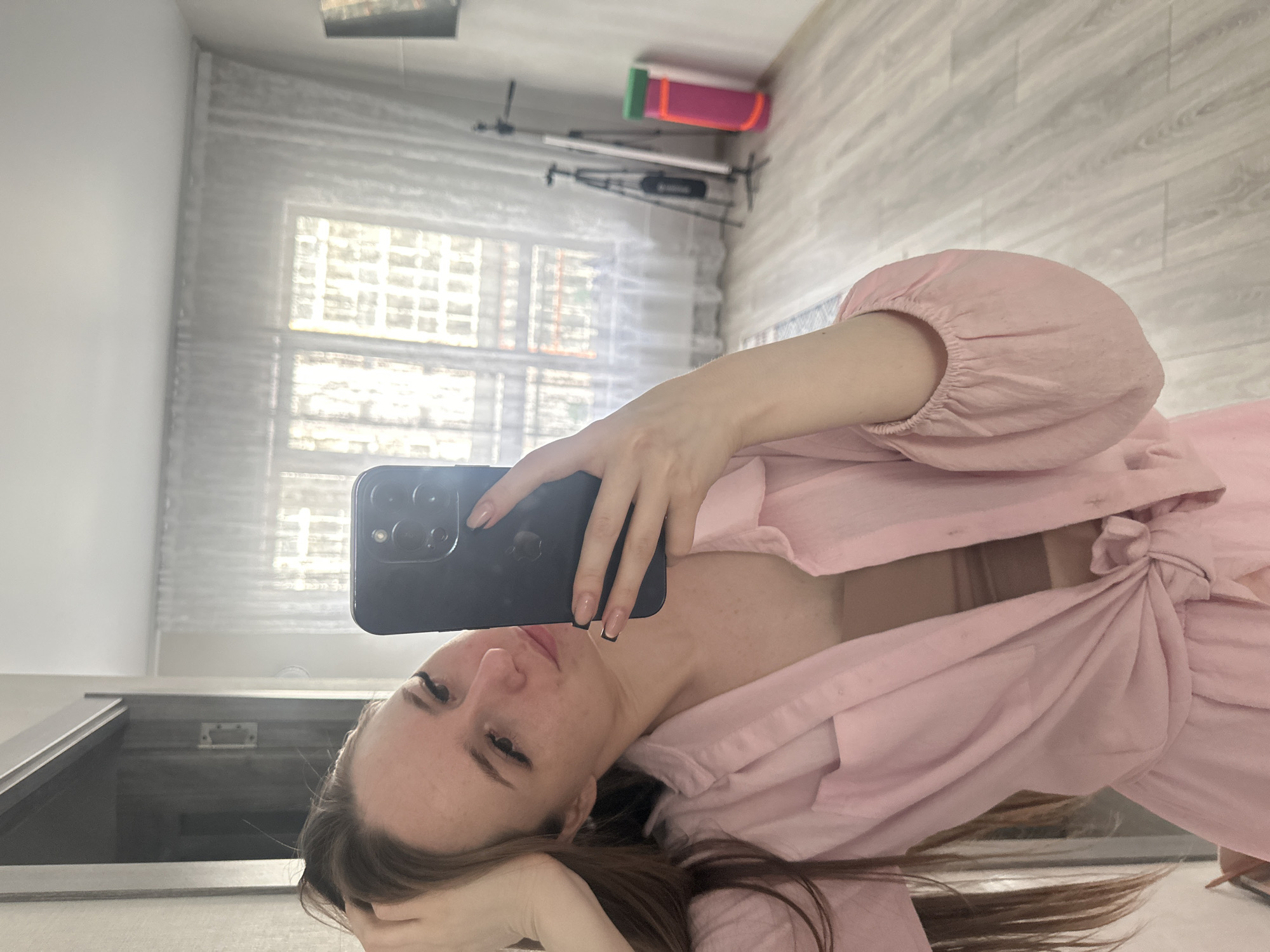 The image shows a mirror selfie of a woman, oriented 90 degrees clockwise. The woman, with long brown hair, appears to be lying down or reclining, with her head resting on her left hand. She is wearing a light pink, long-sleeved shirt or blouse that is tied at the front, revealing a light brown or nude-colored bra underneath. Her eyelashes are prominent, and her nails are manicured with French tips featuring a black outline. She holds a black smartphone, likely an iPhone given the visible Apple logo on its back and camera configuration, up towards a mirror to capture the selfie.
The background shows an indoor setting, likely a room. A large window with horizontal blinds or sheer curtains allows natural light into the space. A light-colored wooden floor covers most of the visible area. In the distance, behind the woman, some equipment resembling tripods or stands, along with a brightly colored pink and green object, are visible. The walls are white. The overall impression is a casual, relaxed moment captured in a residential environment during daylight hours.