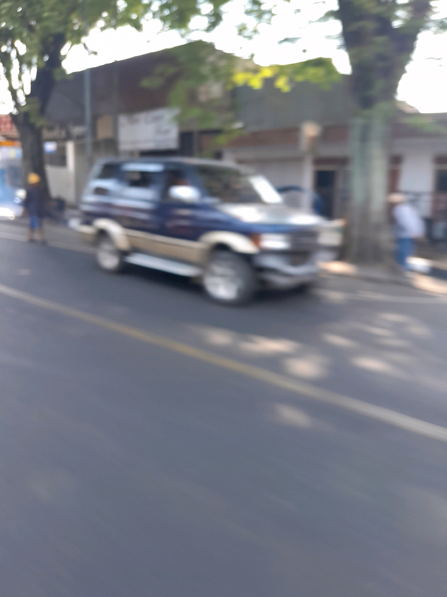 The image depicts a street scene in Kepanjen, Indonesia, with a blue and tan SUV parked on the side of the road. The vehicle is a four-door SUV with prominent wheel wells and what appears to be a roof rack. A person in a dark jacket and blue shorts stands near the front of the vehicle, facing away from the camera. Another person, wearing a light-colored shirt and dark pants, is visible further down the street, also facing away. The background shows buildings with windows and a doorway, characteristic of urban architecture. Trees with green foliage are present on the left side of the image, casting dappled shadows on the road and pavement. The overall lighting suggests it is daytime.