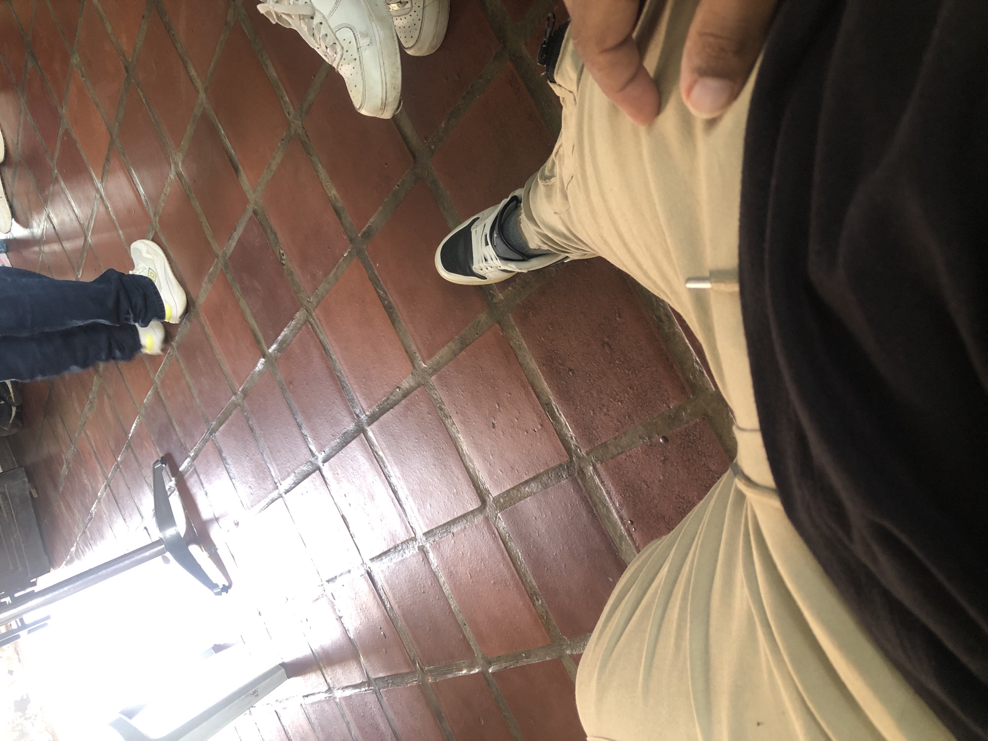The image depicts an indoor scene with tiled flooring. Three pairs of shoes are visible: two pairs of white sneakers and one pair of black and white sneakers. The person wearing the black and white sneakers is also wearing khaki pants and a black shirt or jacket. In the bottom left corner, a metal object resembling a chair or piece of equipment is partially visible. The image does not provide enough information to determine the exact location within El Valle del Espíritu Santo, Venezuela, or the actions and interactions occurring in the scene.
