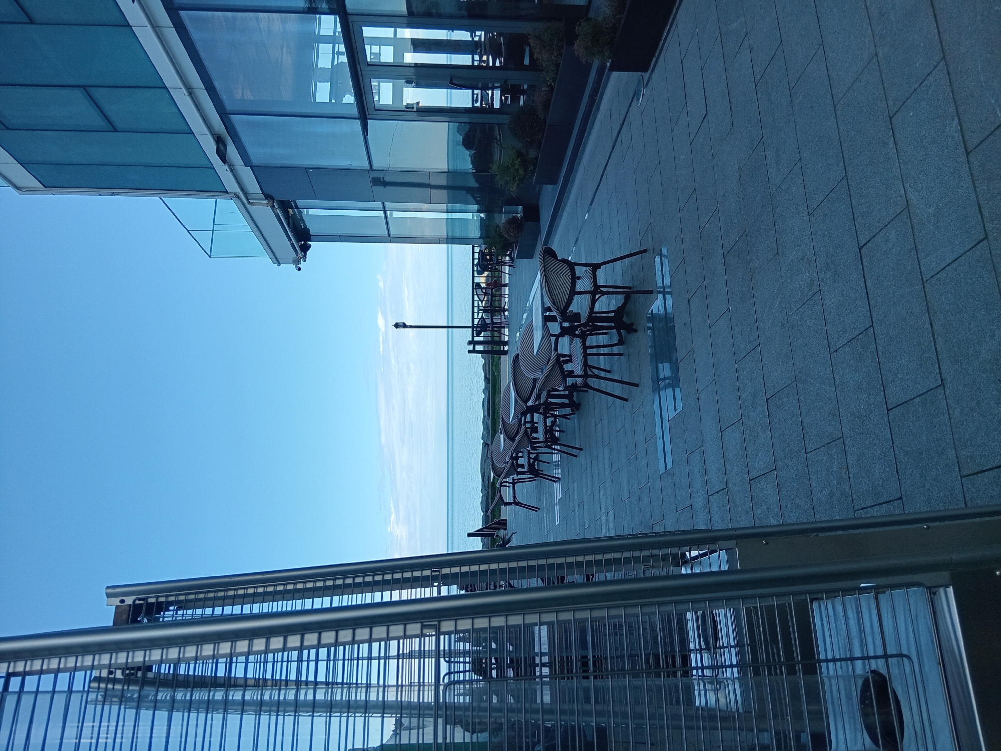 The image depicts a waterfront area in Batumi, Georgia. The scene captures outdoor seating with chairs and tables set on a tiled patio next to a building with large glass windows, reflecting the sky and sea. A metal railing is visible in the foreground. The Black Sea stretches to the horizon under a clear, blue sky.
