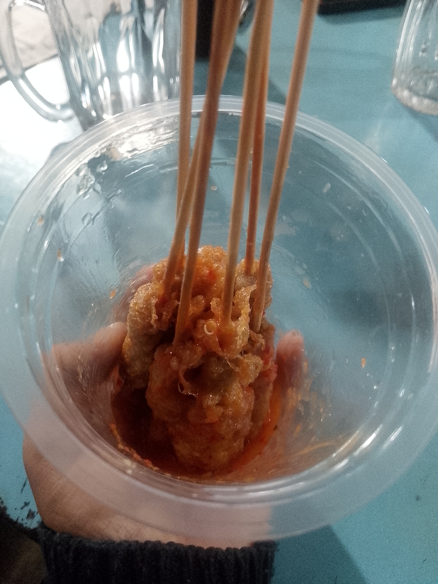 A close-up shot shows a hand holding a clear plastic cup filled with food on skewers. The food appears to be small pieces of meat covered in a reddish-orange sauce. The skewers are made of light-colored wood. In the background, out of focus, are elements that suggest an urban environment, possibly a street food stall in Lhokseumawe, Indonesia, with a glass in the upper left and a blue surface that could be a table. The lighting is somewhat dim, with reflections on the plastic cup.